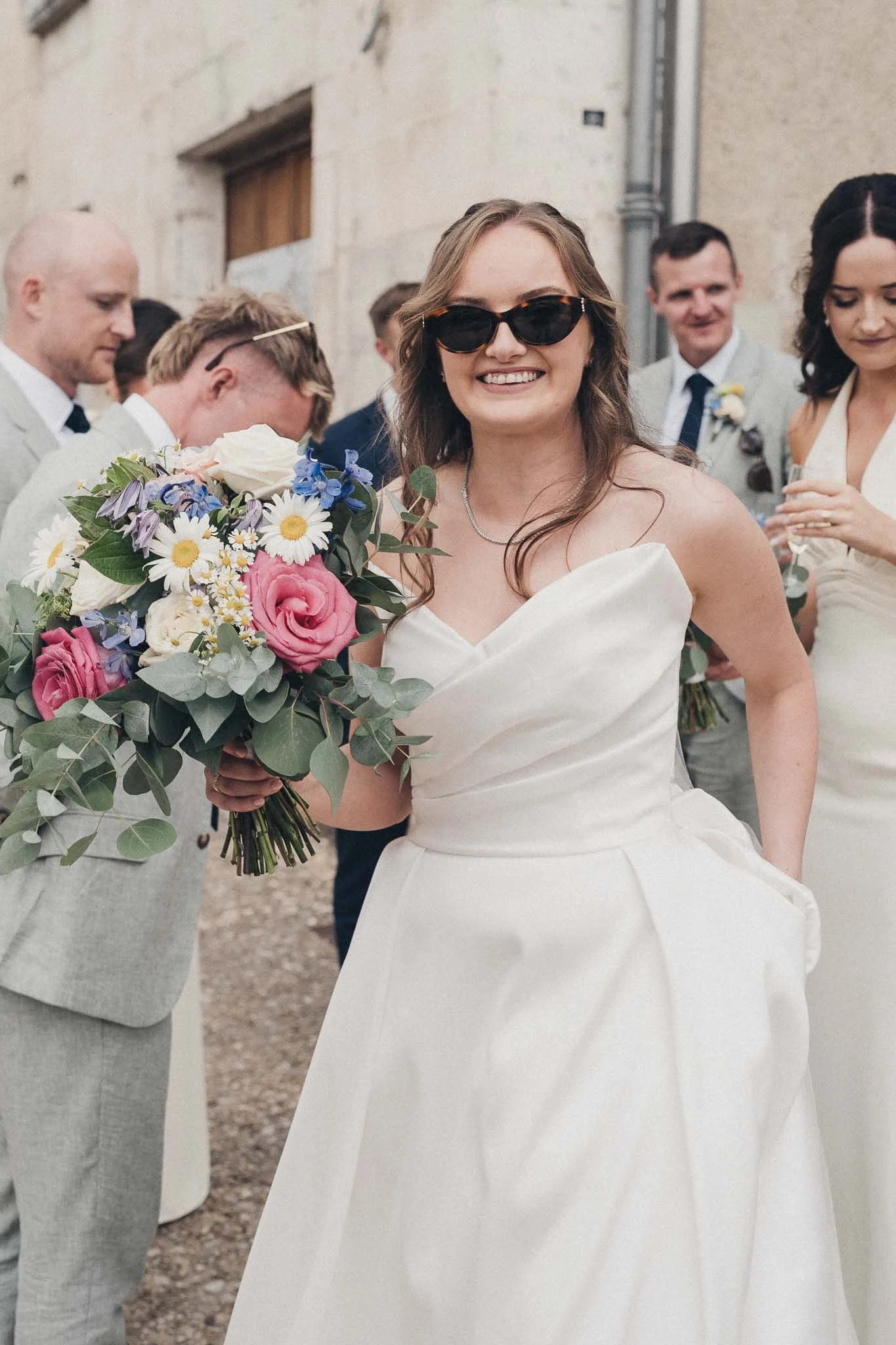 Bride smiling with sunglasses on, holding her bouquet during the celebrations.
