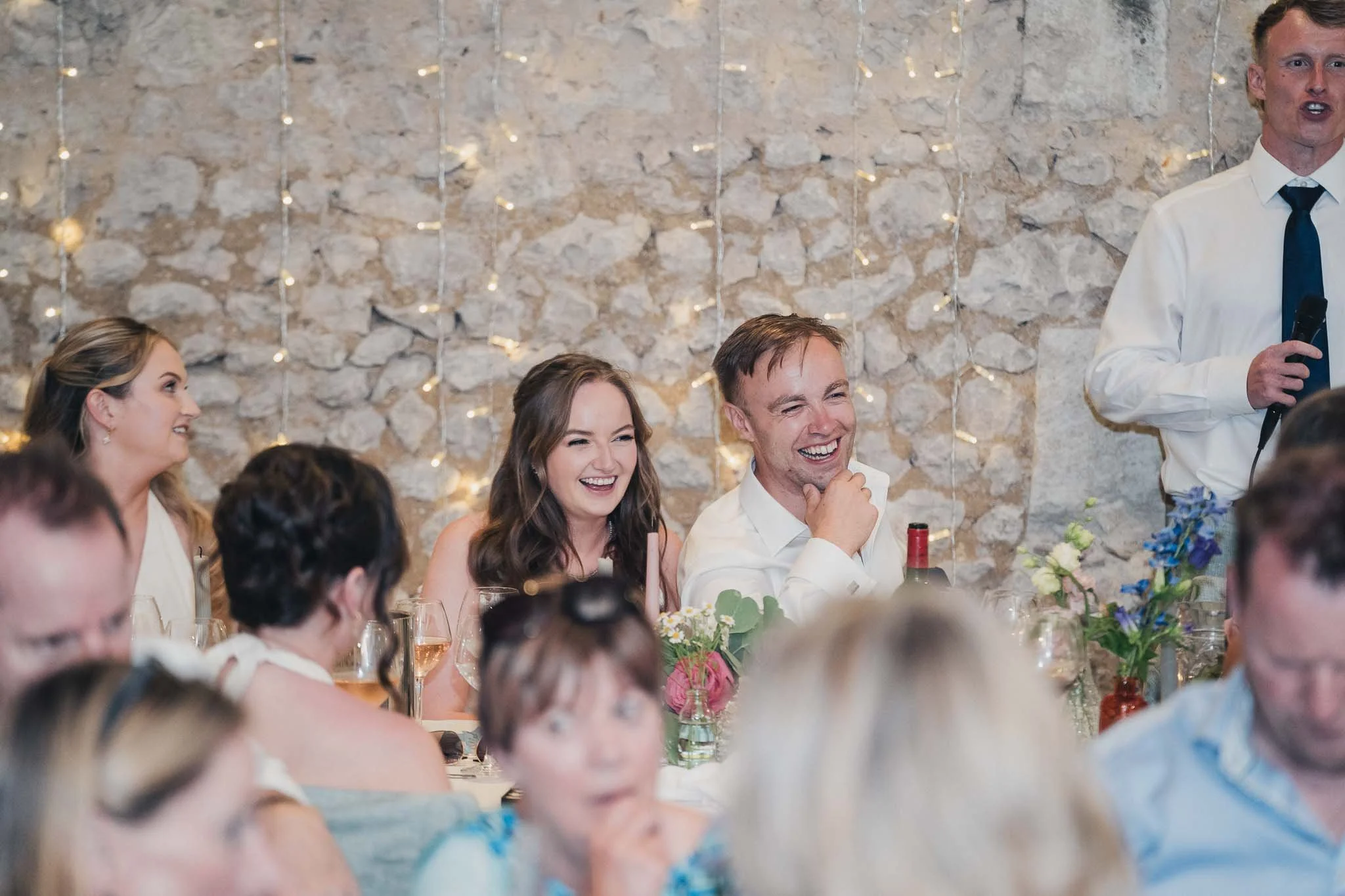 Bride and groom smiling and reacting during a wedding speech at dinner.
