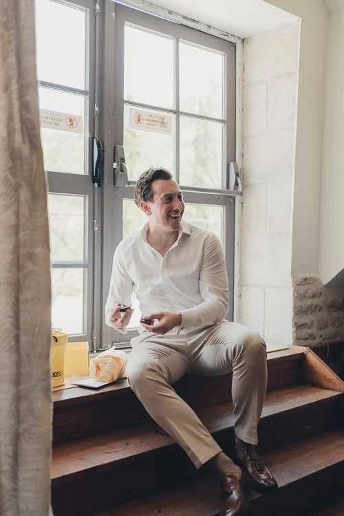 Groom sitting on stone steps during the wedding day, laughing quietly with his groomsmen in an unguarded moment between events.