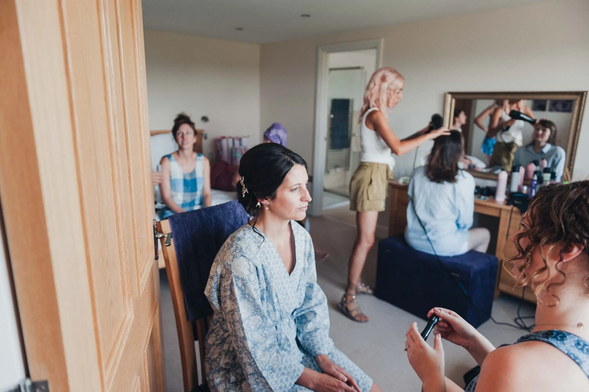 The bride sitting for hair and makeup while the room buzzes with people getting ready.