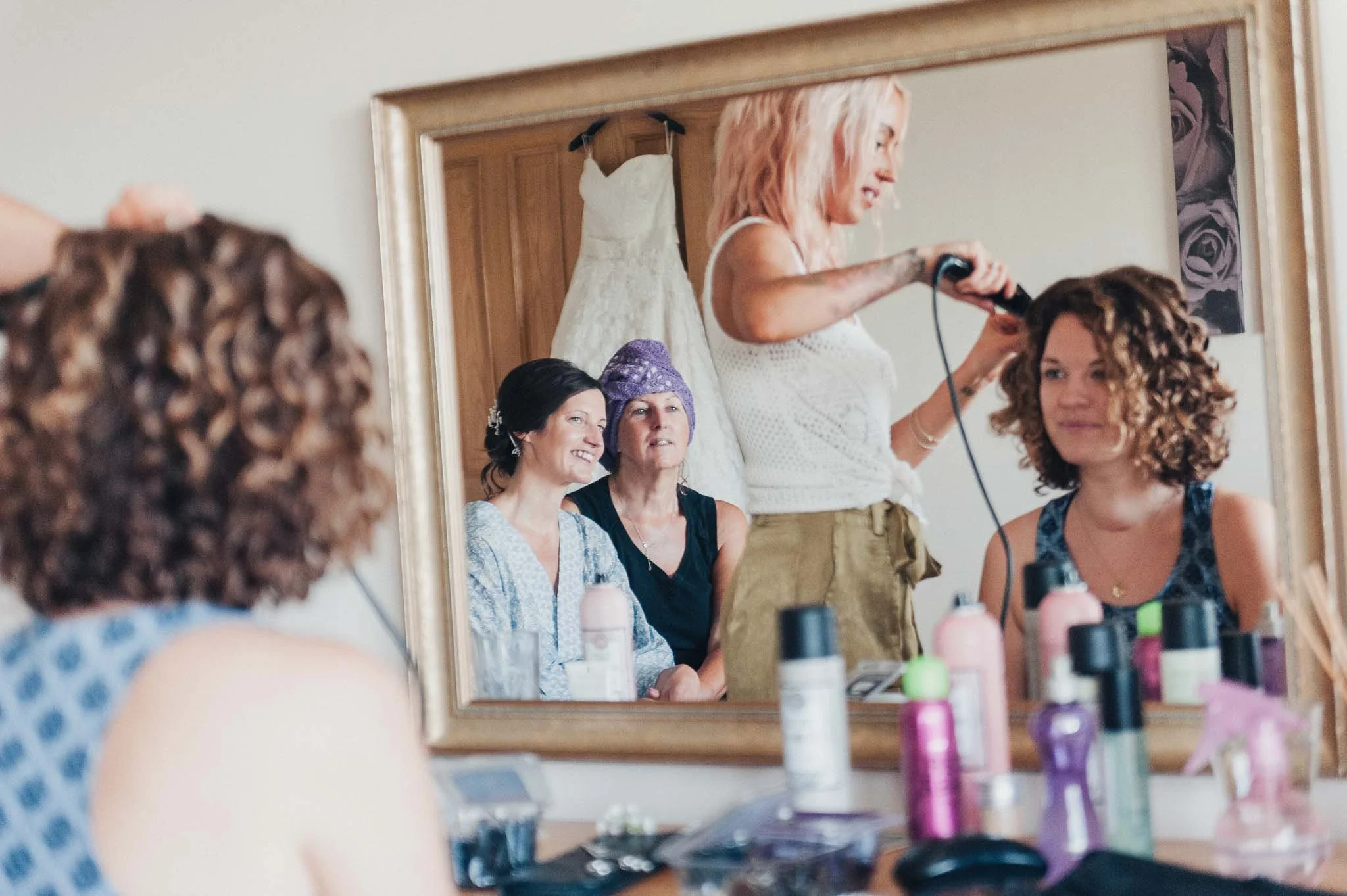 A bridesmaid having her hair styled, reflected in a large mirror with the bride and her mum in the background.