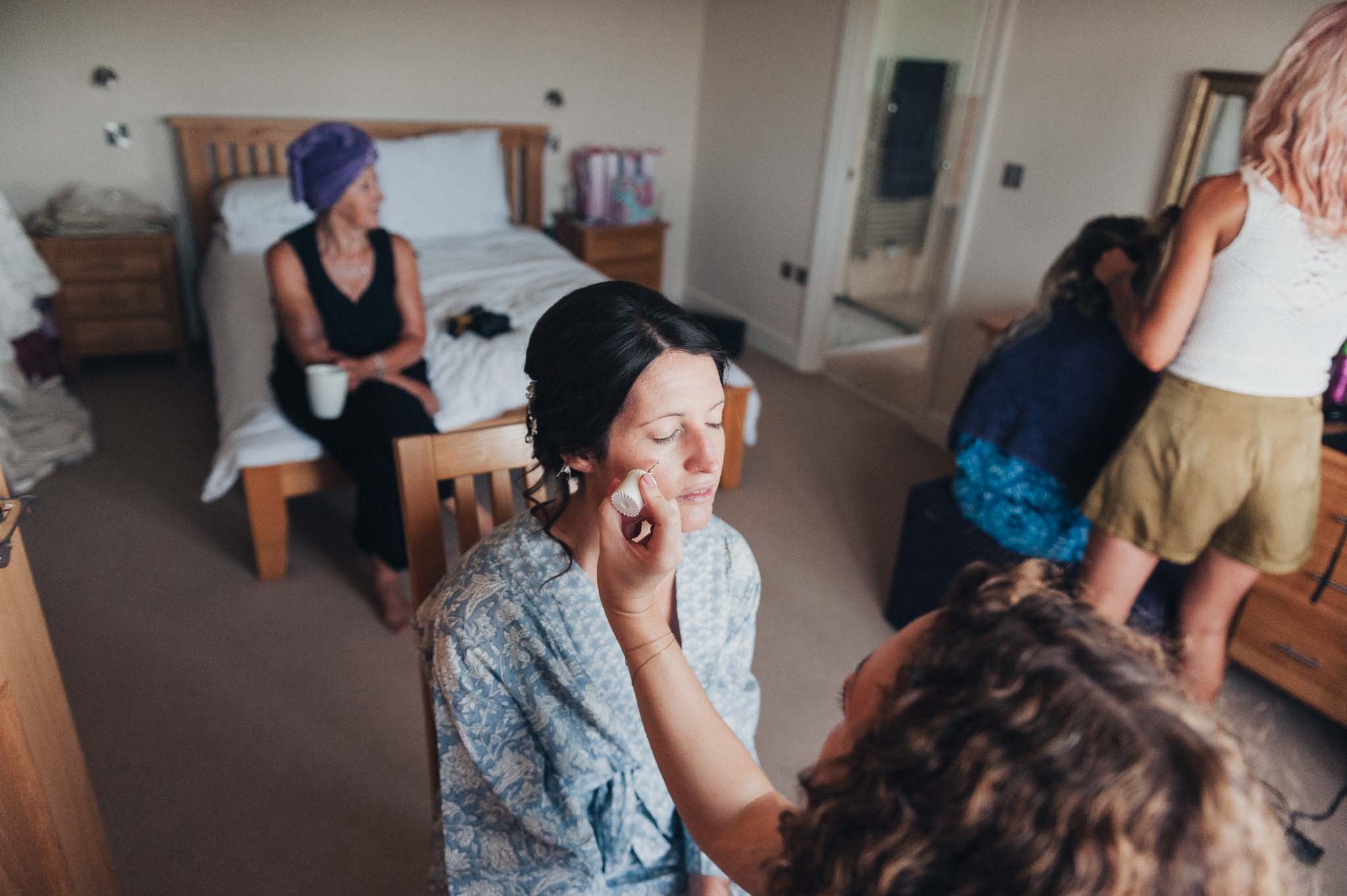 The bride having her makeup done in a bright bedroom while bridesmaids get ready around her.