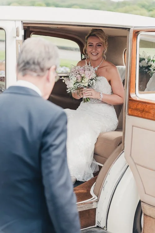 Bride stepping out of a vintage wedding car with her father just before the ceremony, a calm moment before walking inside.