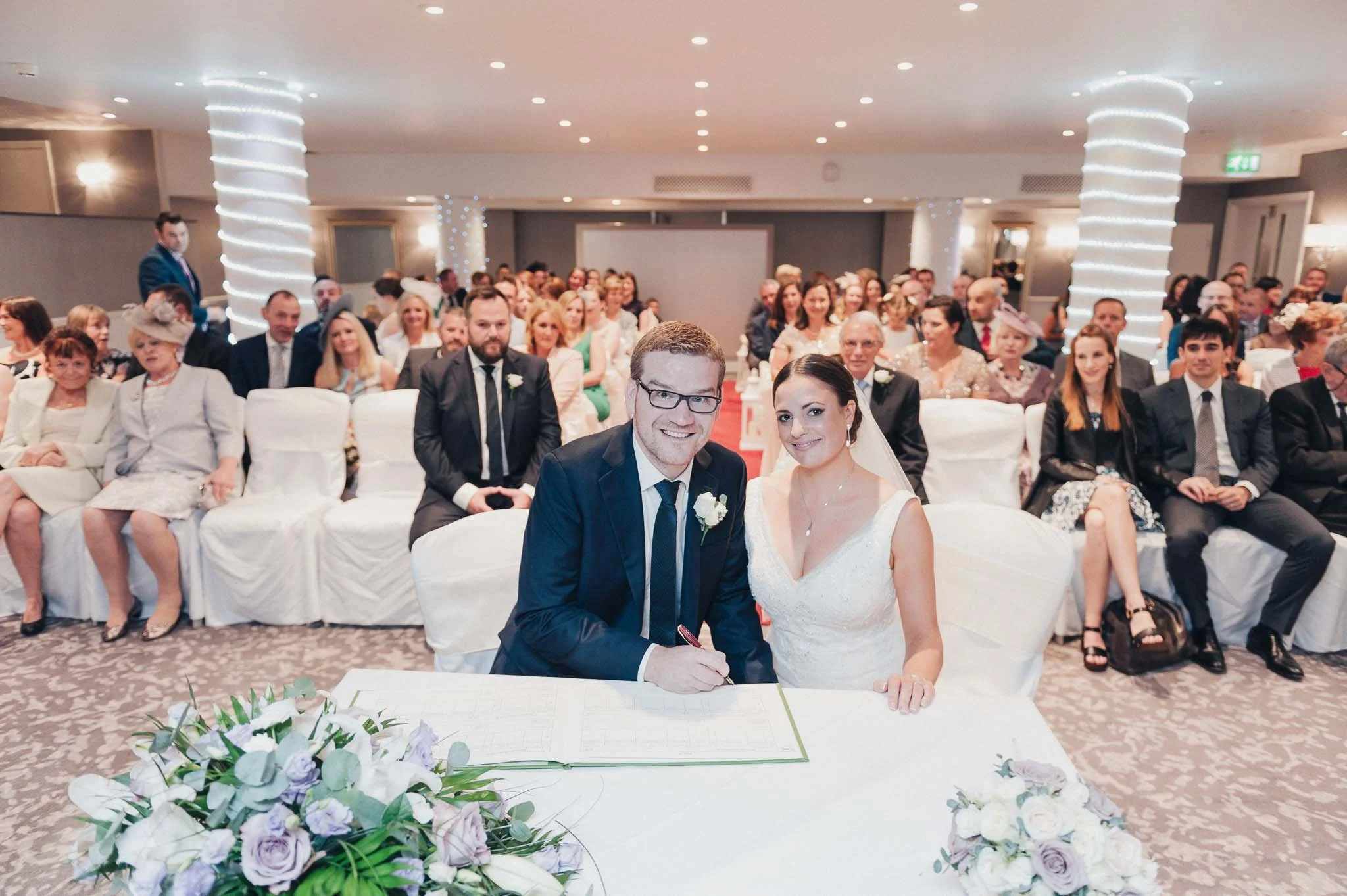 Bride and groom signing the marriage register in front of their guests during the indoor ceremony.