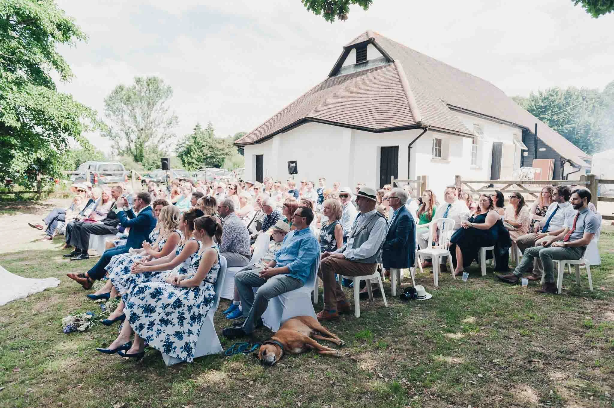 Wide view of the outdoor ceremony with guests seated facing the couple and the village hall in the background.