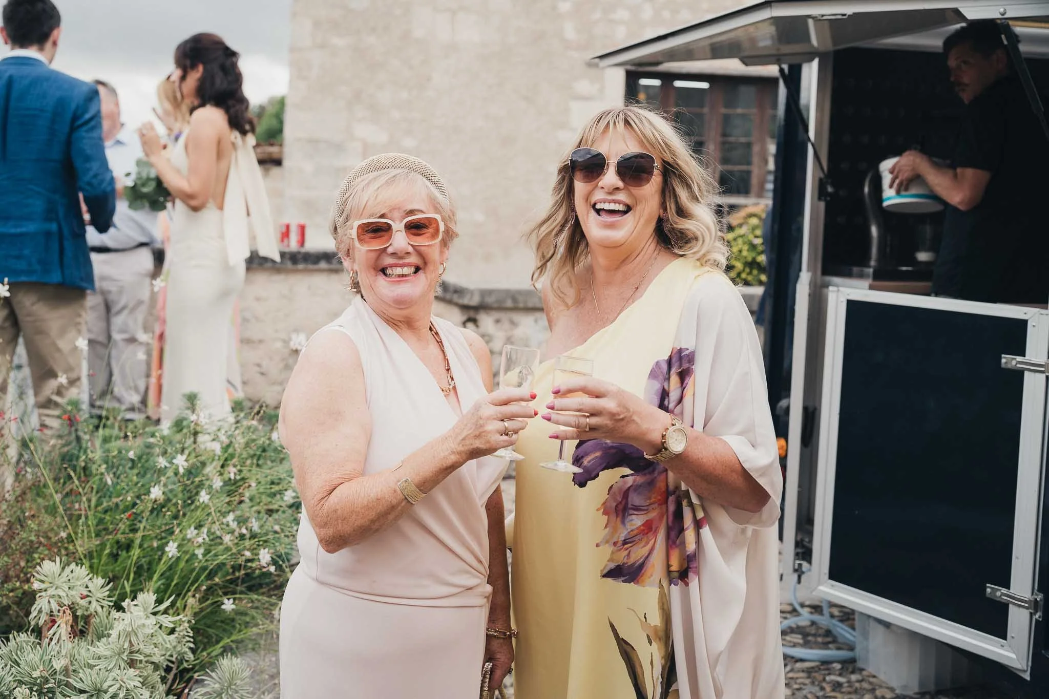 Two guests smiling and holding drinks during the outdoor reception at Manoir de Longeveau.