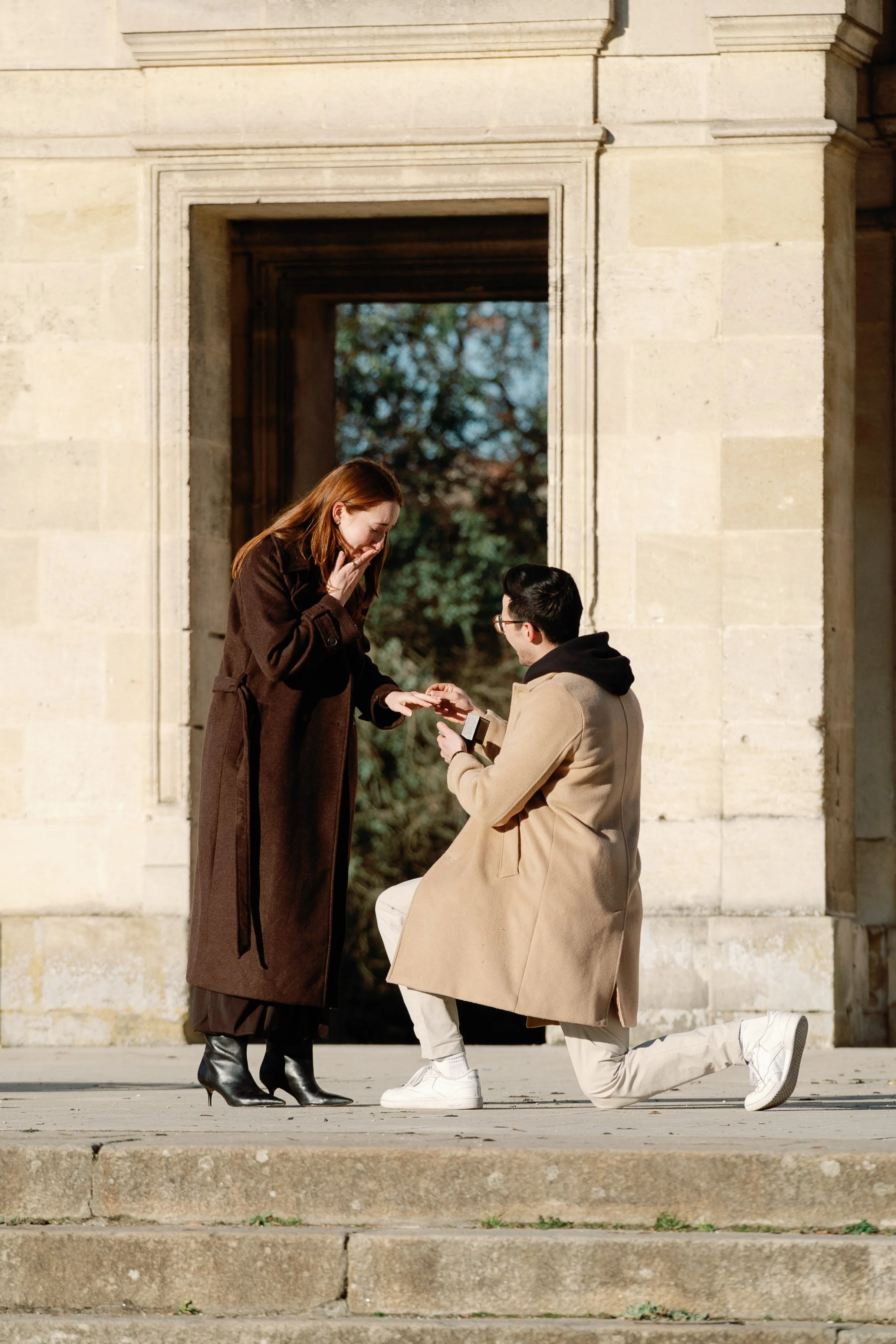 Man proposing on one knee beneath stone arches in Bordeaux as his partner reacts emotionally.