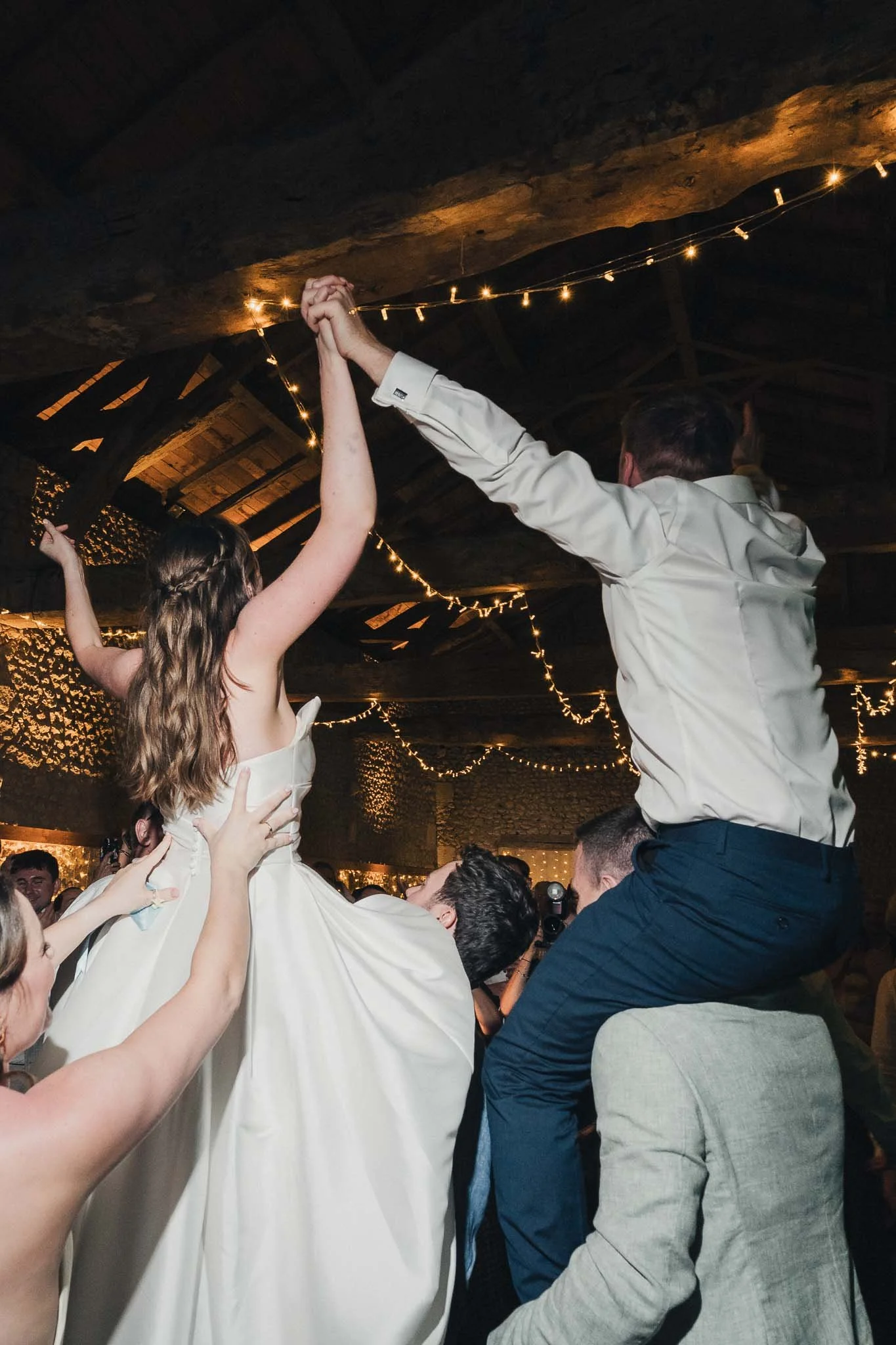 Bride being lifted up by guests with her arms raised on the dancefloor inside the barn.