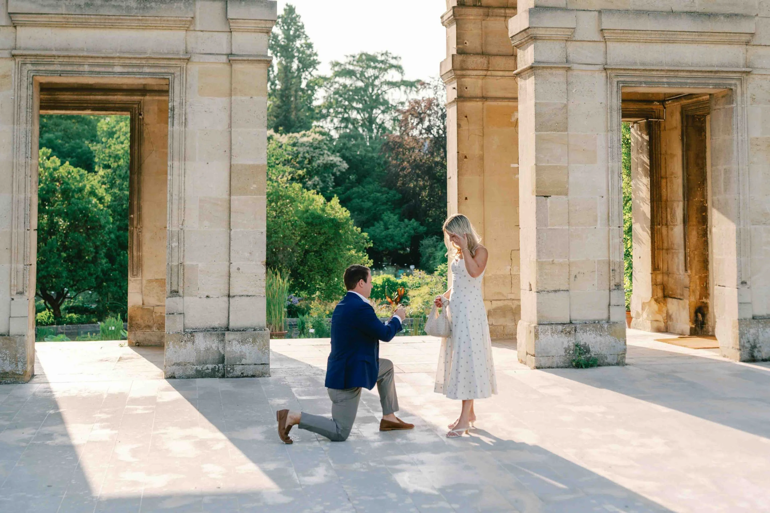 Man on one knee proposing between the stone columns of the Palais in Jardin Public, Bordeaux, as she covers her face in surprise
