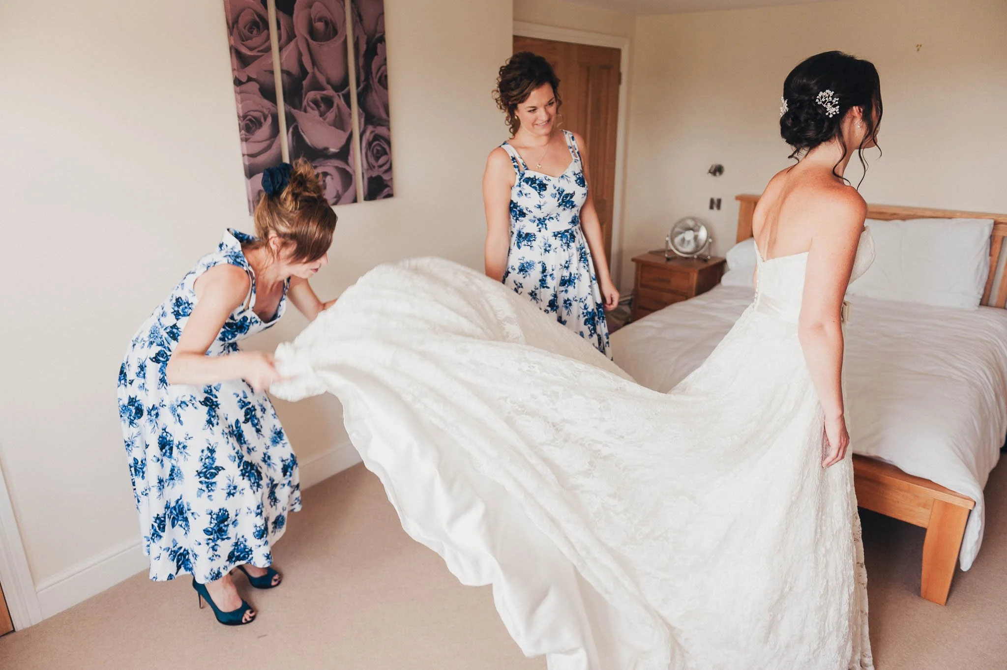 A bridesmaid lifting the train of the bride’s wedding dress while getting ready in the bedroom.