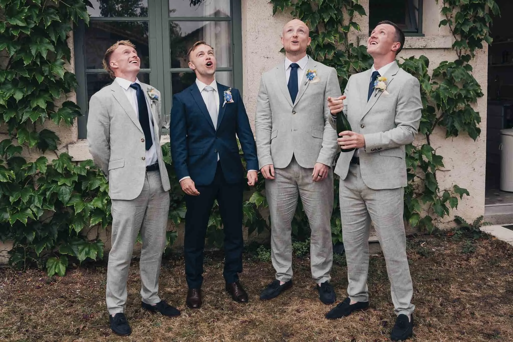 Groom and groomsmen laughing while opening a champagne bottle outside Manoir de Longeveau.