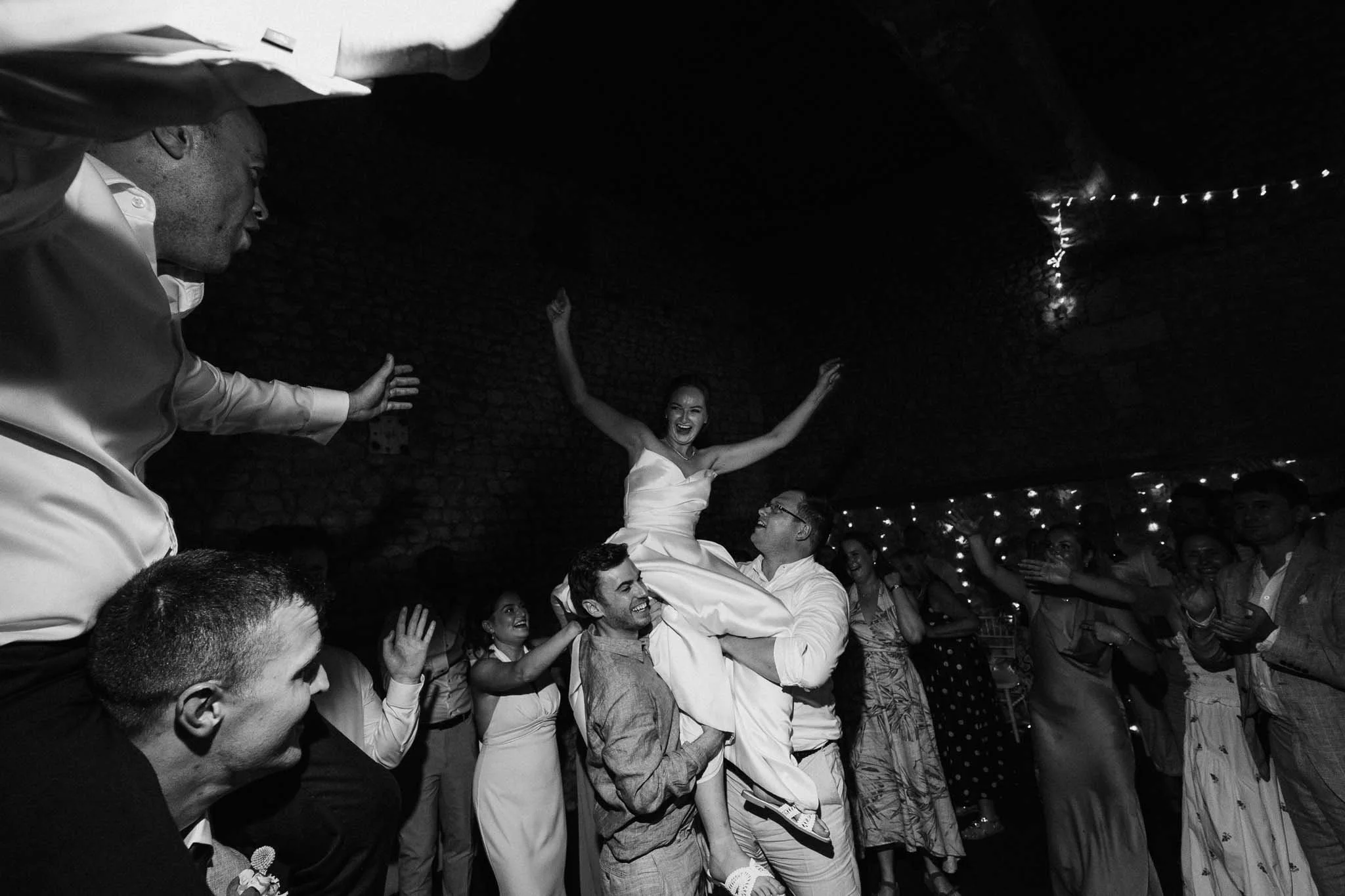 Black-and-white photo of the bride being lifted into the air on the dancefloor while guests celebrate around her.