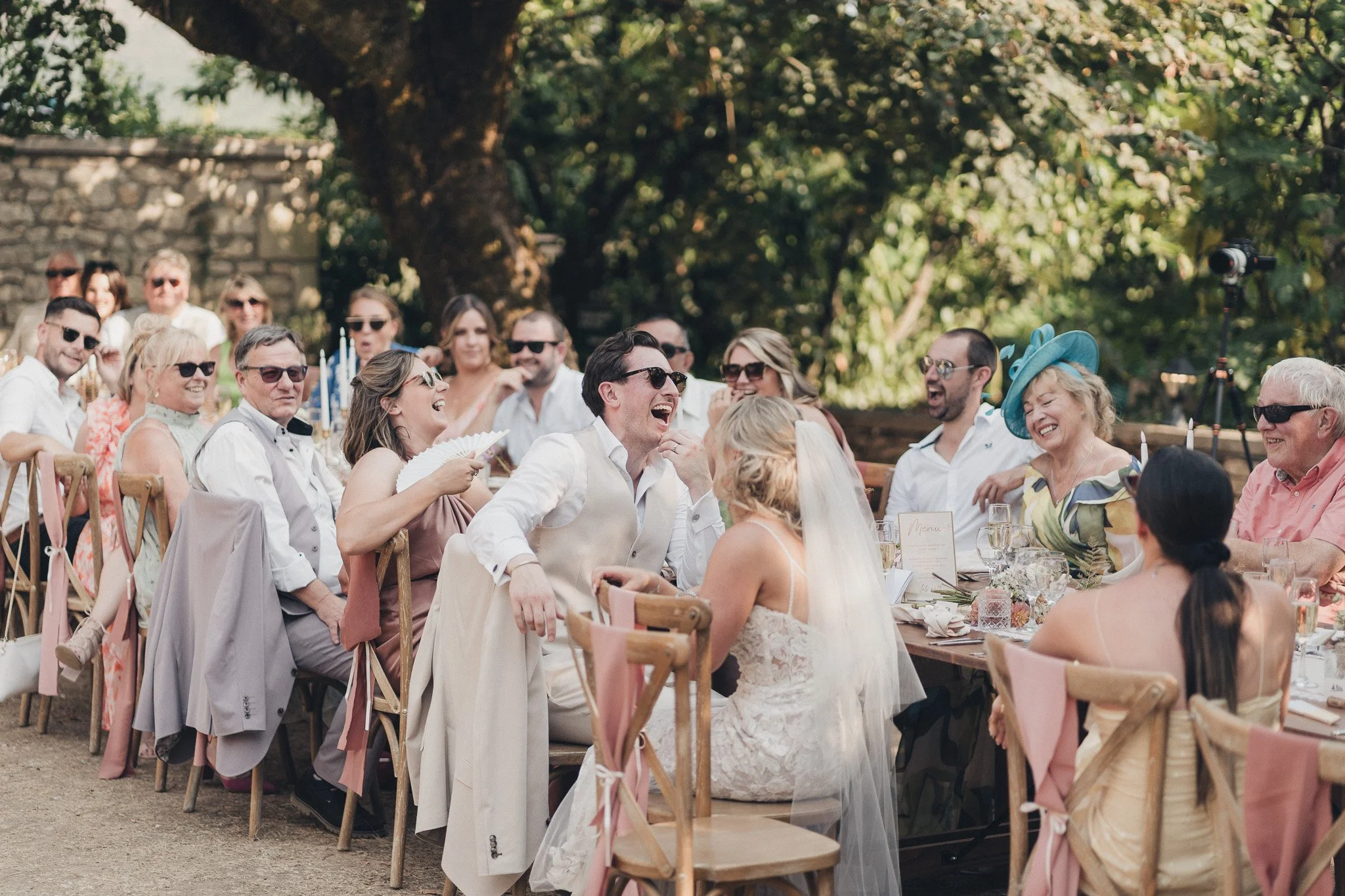 Guests laughing during outdoor wedding speeches at a long table reception in southwest France.