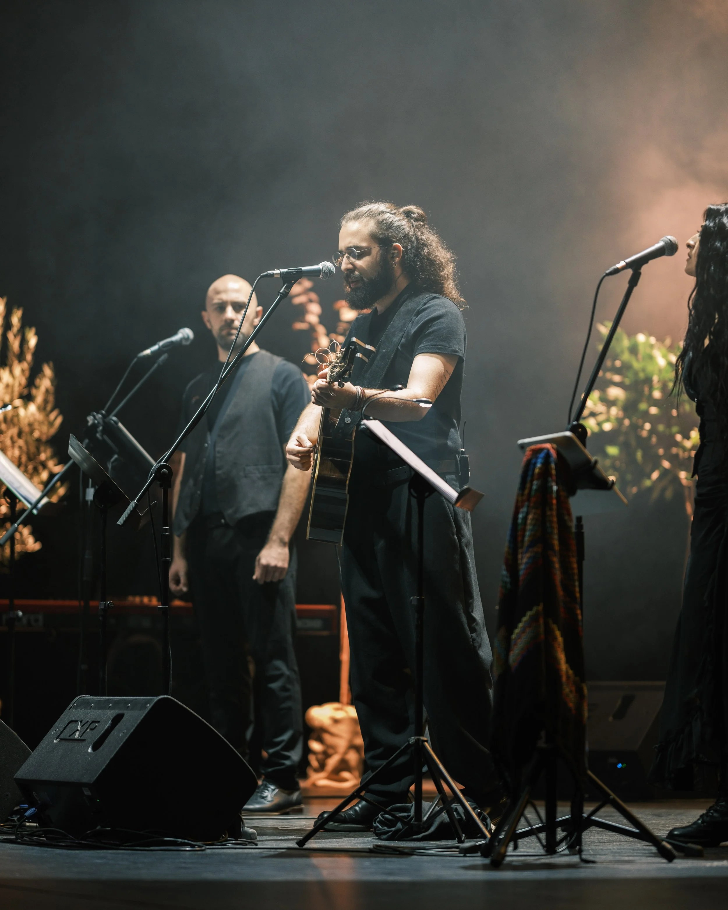 Musicians performing on stage, including a man with long curly hair playing guitar and singing into a microphone, with two other musicians standing nearby, surrounded by musical equipment and stage lighting.