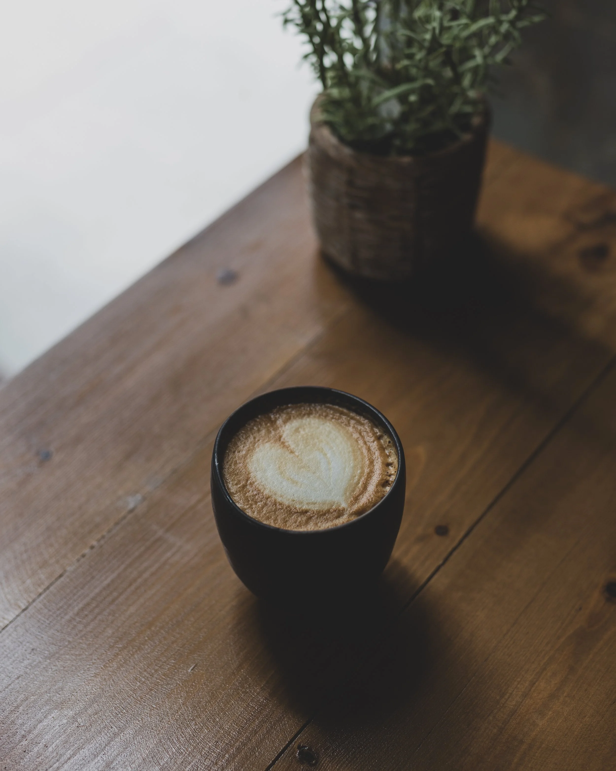 A black cup of coffee with frothy milk and a heart-shaped latte art on a wooden table next to a potted plant.
