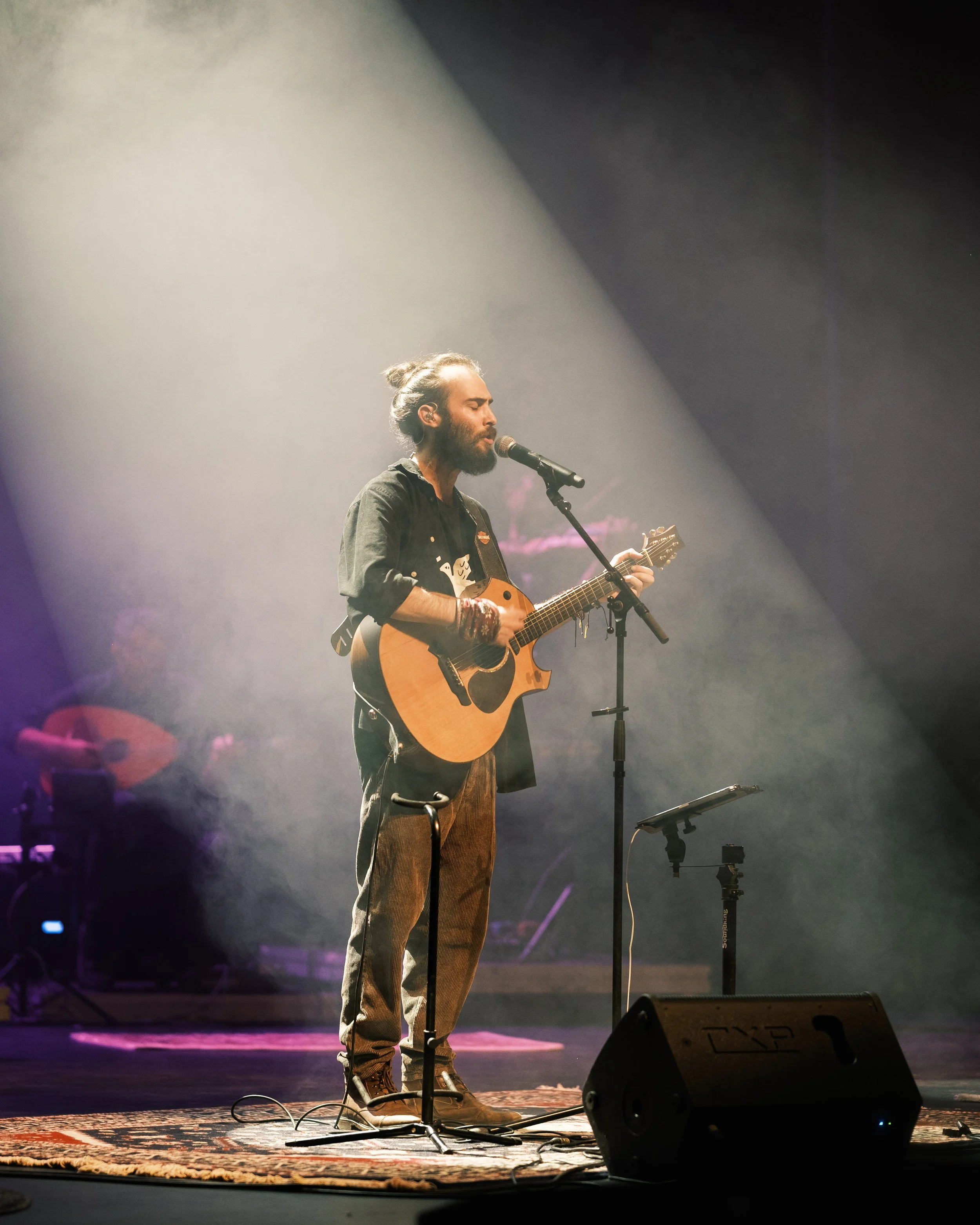 A man with long hair tied in a bun, beard, wearing a dark shirt, brown pants, and boots, is performing alone on stage with an acoustic guitar, singing into a microphone, with stage lights and equipment visible in the background.