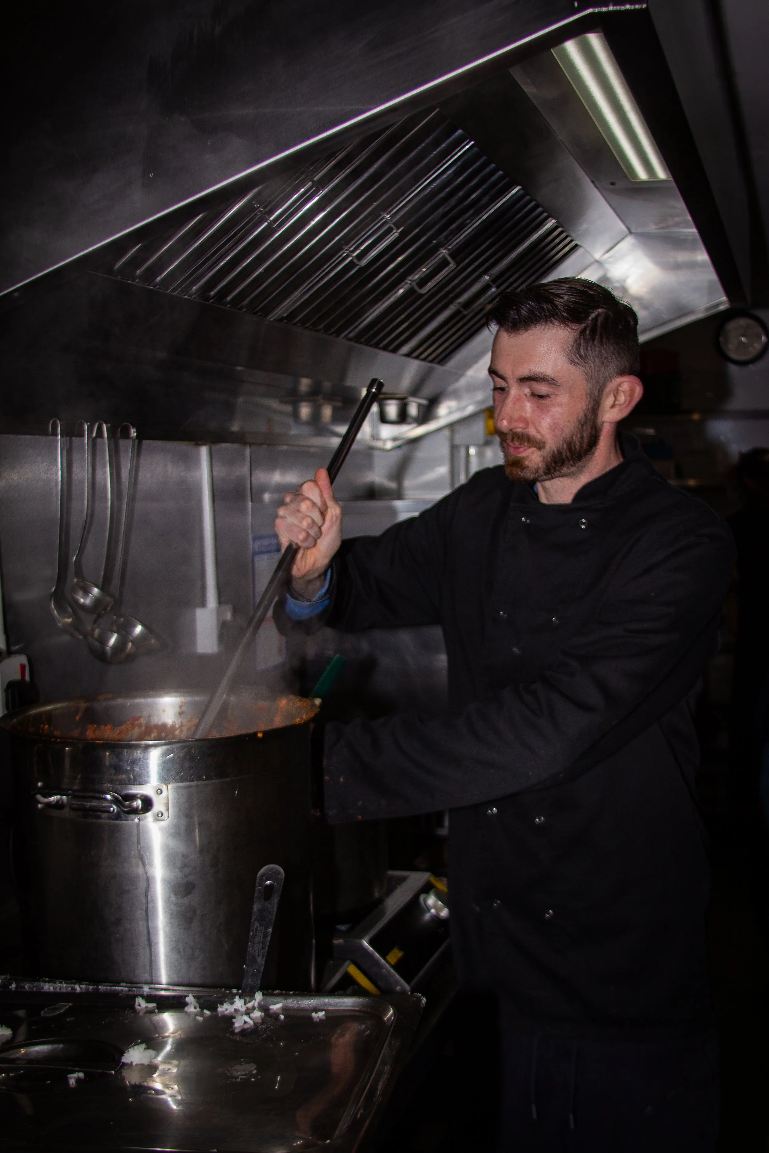 A man in a black chef jacket stirs a large pot of pasta sauce in a professional kitchen.