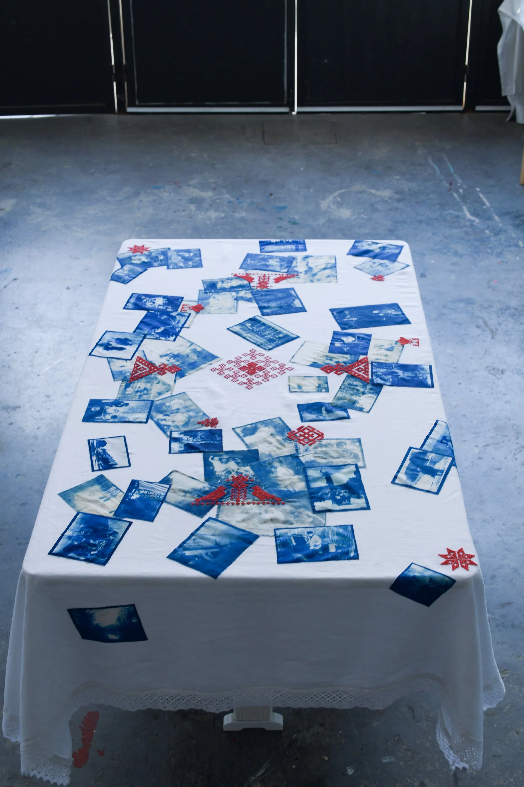 A table covered with an embroidered white tablecloth decorated with multiple blue photos and red embroidery designs, situated on a concrete floor in a room with black cabinets in the background.