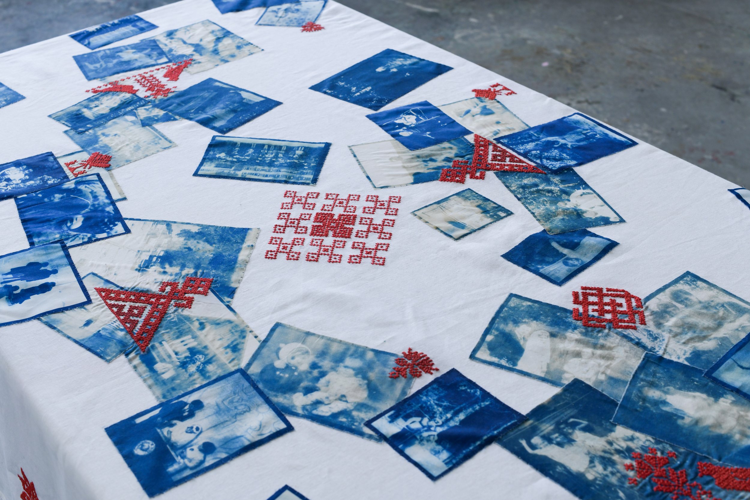 A white cloth with blue and red cross-stitch patterns and printed photographs of snowy mountain scenes and winter landscapes. The cloth is laid out on a surface outdoors.