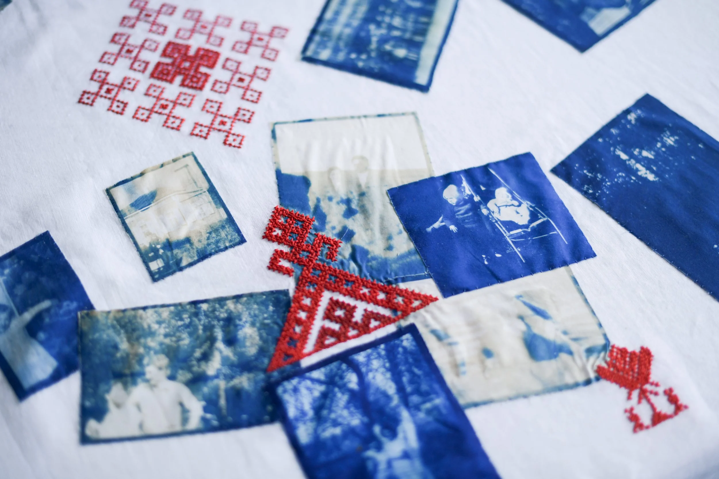 Close-up of embroidered red and blue cross-stitch patterns on white fabric, including small landscape photographs featuring people and outdoor scenes.