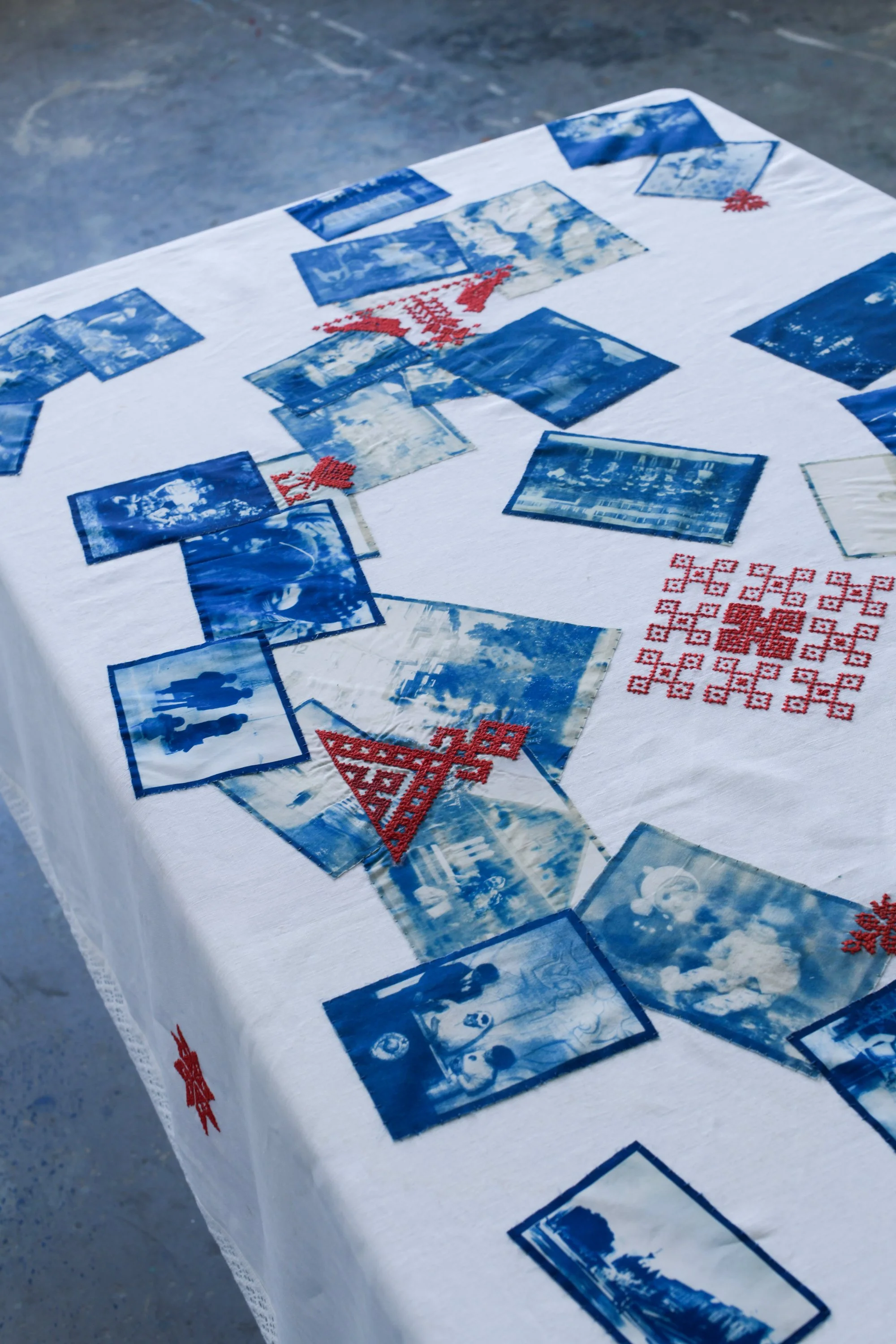 A table covered with a white cloth displaying numerous blue and white photographic squares and red embroidered patterns, with a gray concrete floor in the background.