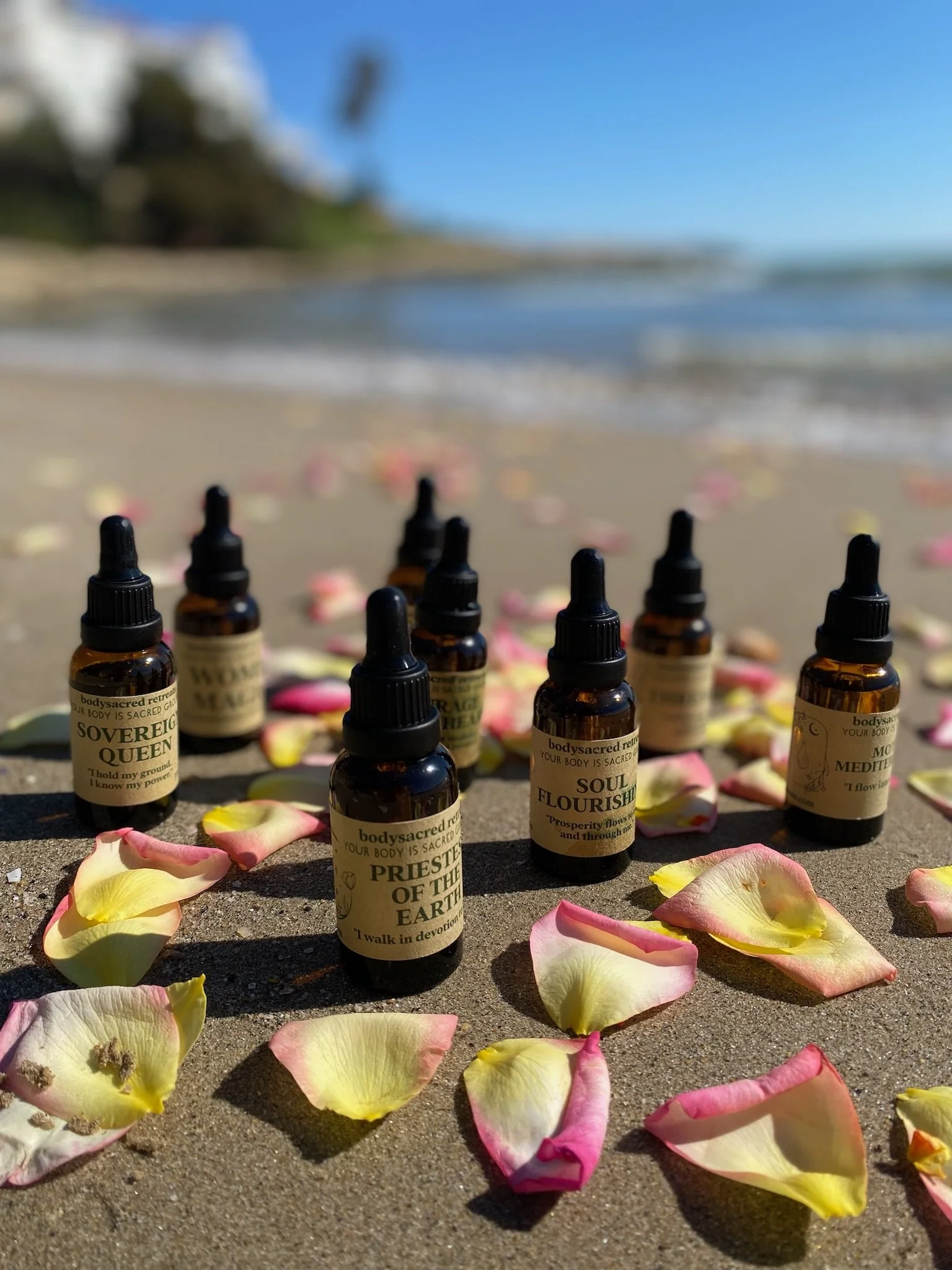 Small brown bottles with labels reading 'Soul Flourish' and 'Priestess of the Earth' placed on sand with pink and yellow flower petals scattered around, near the water's edge. The background shows a blurred shoreline with trees and a blue sky.