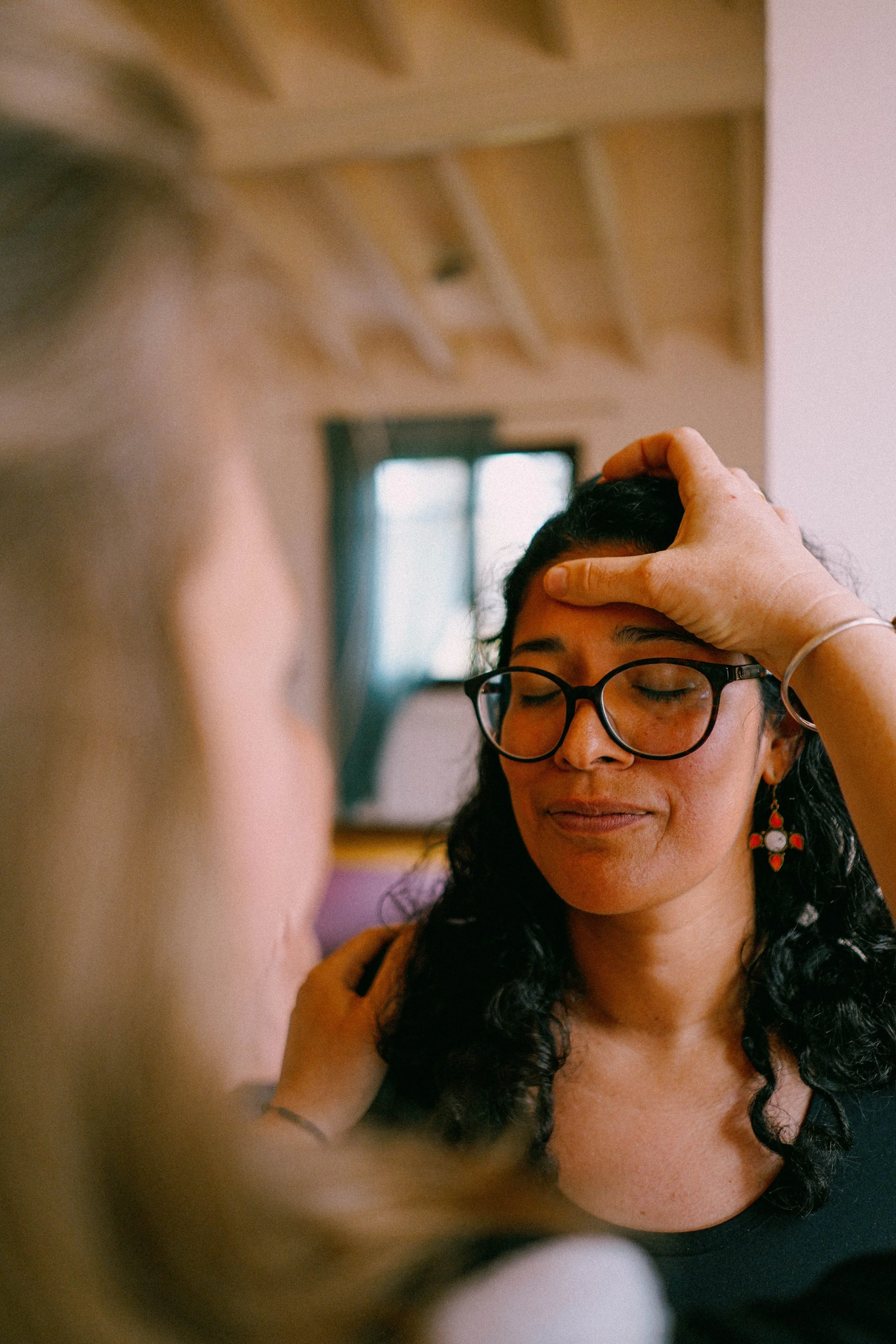 A woman with curly black hair, glasses, and earrings, gently touching her forehead, appears to be in a calming or meditative moment.