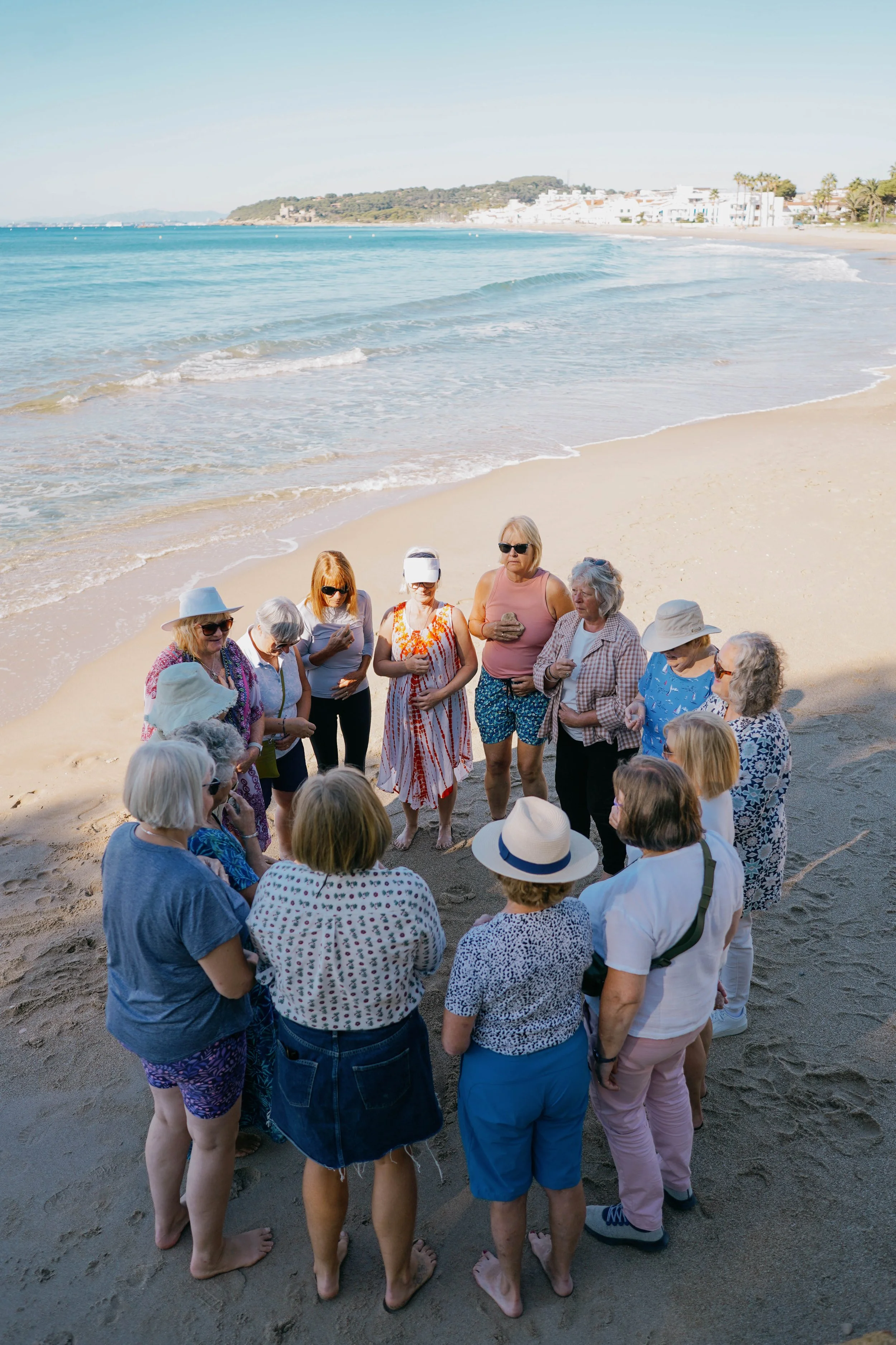 A group of older adults standing on a sandy beach in a circle, some wearing sunglasses and summer clothing, with the ocean and buildings in the background.