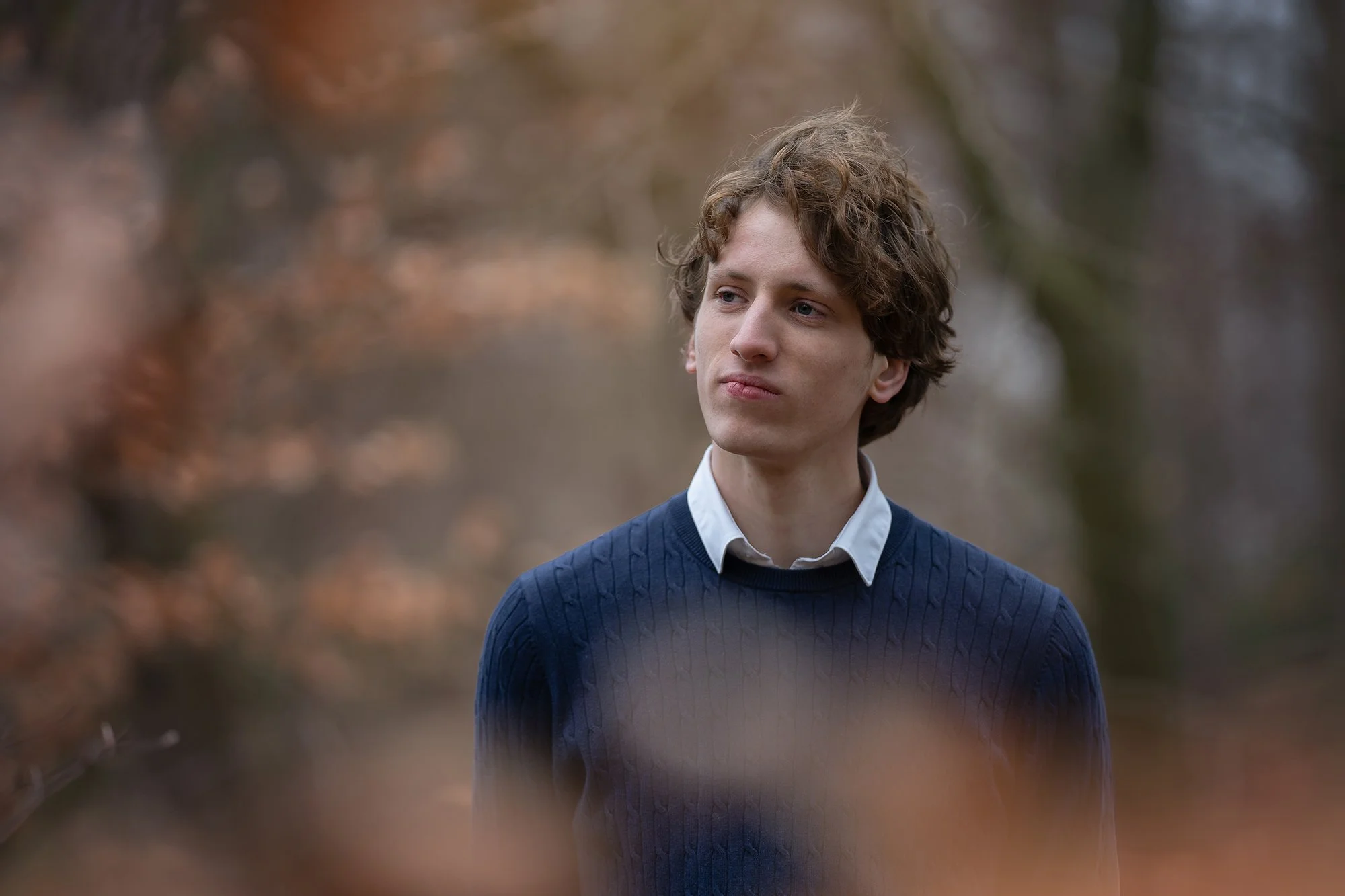 A young man with curly hair wearing a blue sweater and white collared shirt, standing outdoors in a blurred autumnal background.