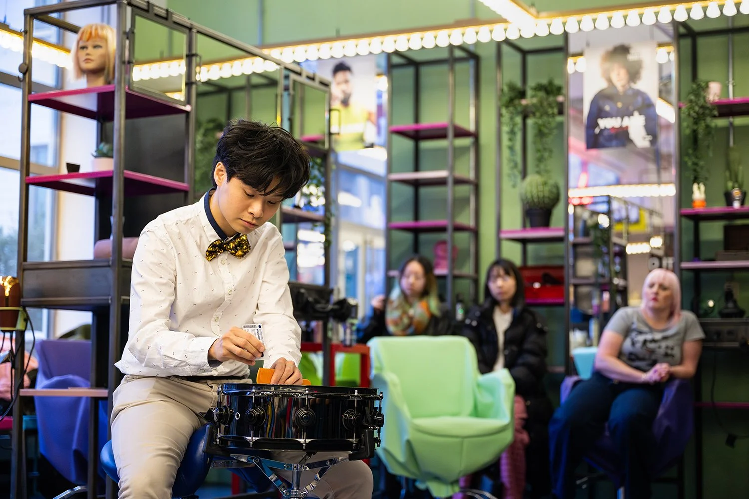 A young man with dark hair wearing a white shirt and a black and yellow bowtie playing a small drums set in a colorful, modern room with shelves, plants, posters, and three women seated in the background.