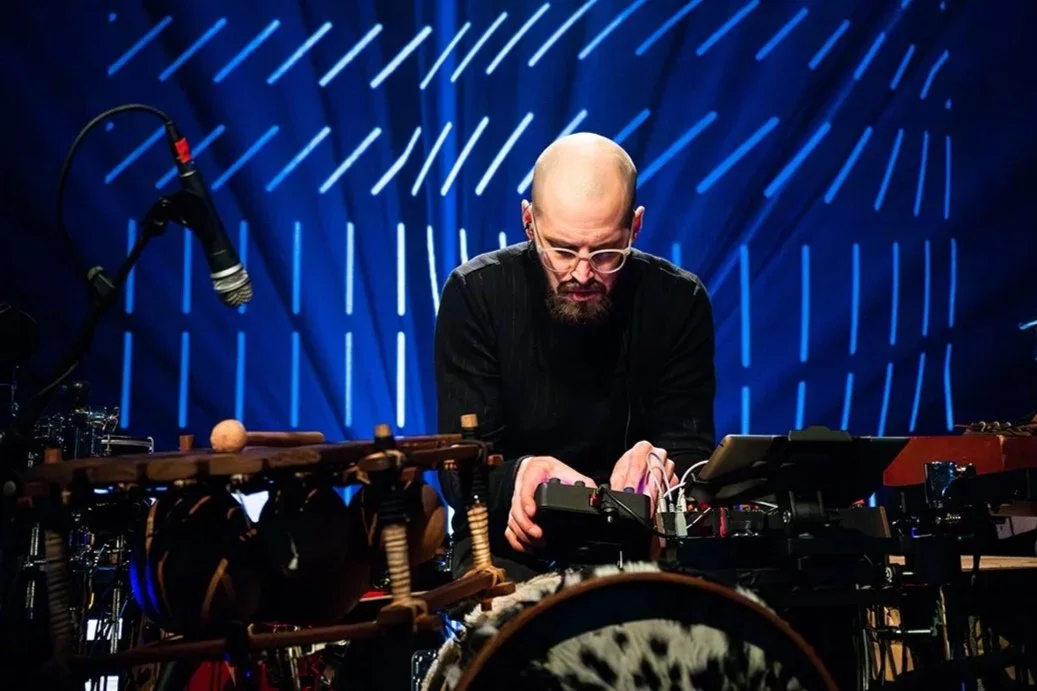 A man with a bald head, glasses, and a beard playing electronic musical instruments during a performance with blue stage lighting and abstract light patterns in the background.