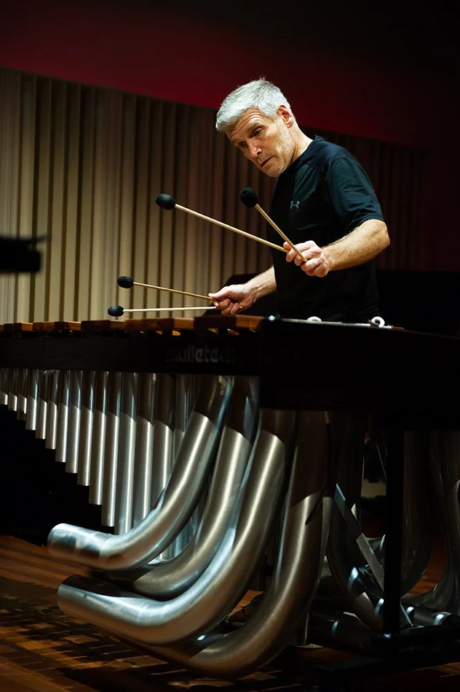 A man with gray hair playing a marimba with four mallets in a music studio or concert hall.