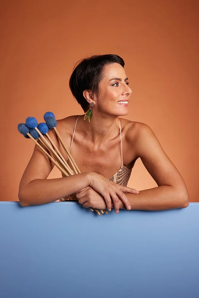 A woman with short dark hair wearing a spaghetti strap top, holding a bunch of blue-tipped knitting needles with her left arm, and smiling while looking to the right, against a warm orange background.