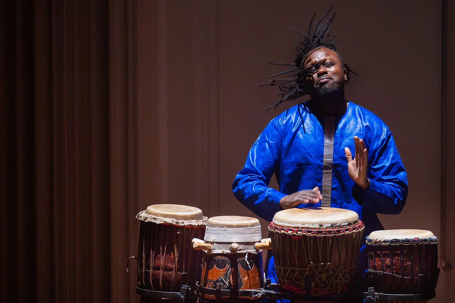 A man playing African drums, wearing a shiny blue traditional outfit, with dreadlocked hair, on stage with a dark background.