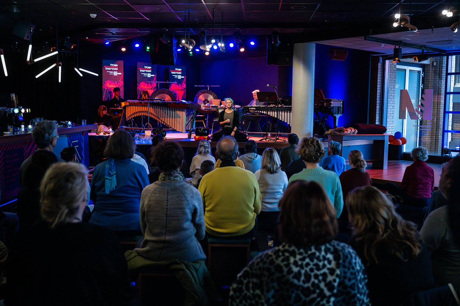 A live music performance featuring a woman singing on stage with children and adults seated in front, in a venue with colorful lighting and musical instruments.