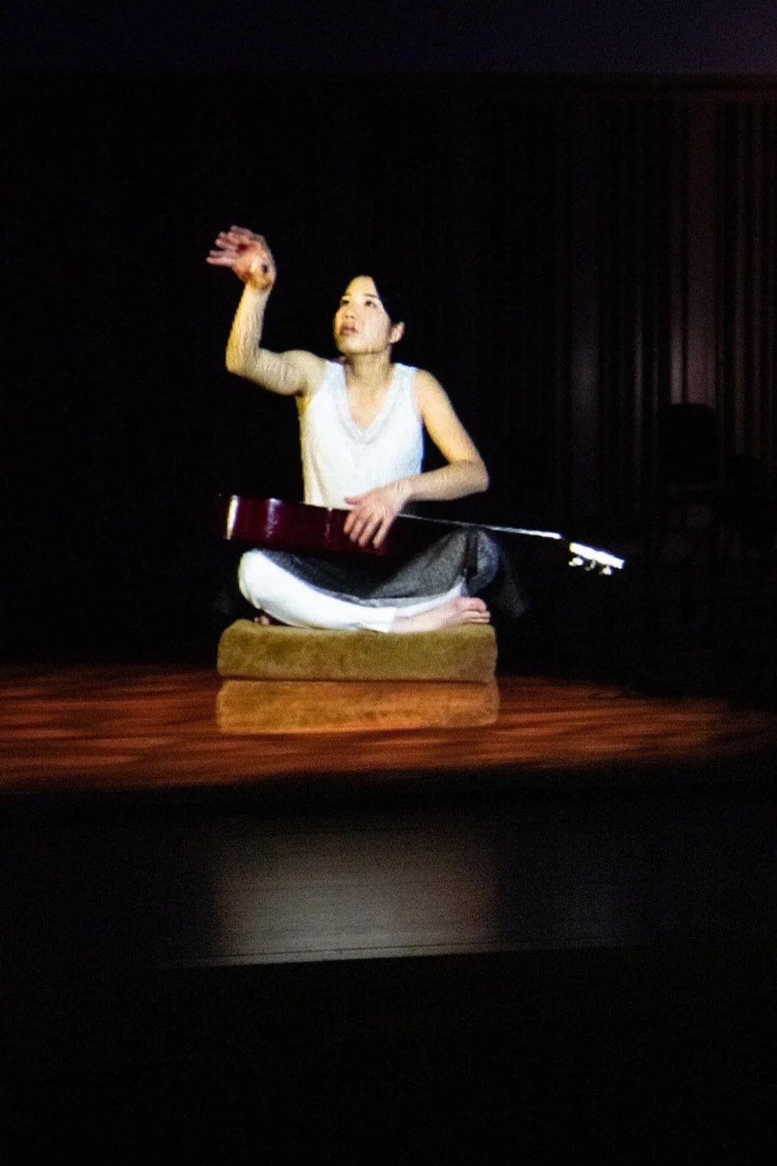 Nozomi Hiwatashi sitting cross-legged on stage with a traditional musical instrument, raising her right hand in a rhythmic gesture, playing as a hologram during the final of Tromp Percussion Eindhoven 2020 competition