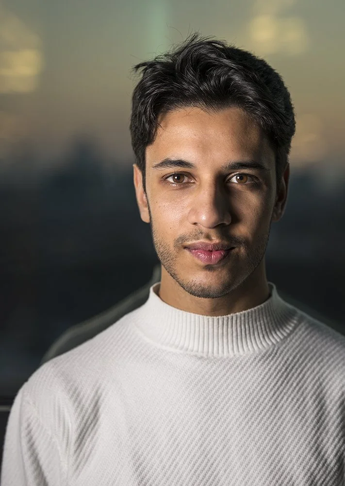 Close-up portrait of a young man with dark hair and brown eyes, wearing a white ribbed turtleneck sweater, standing outdoors with a blurred sunset or sunrise background.