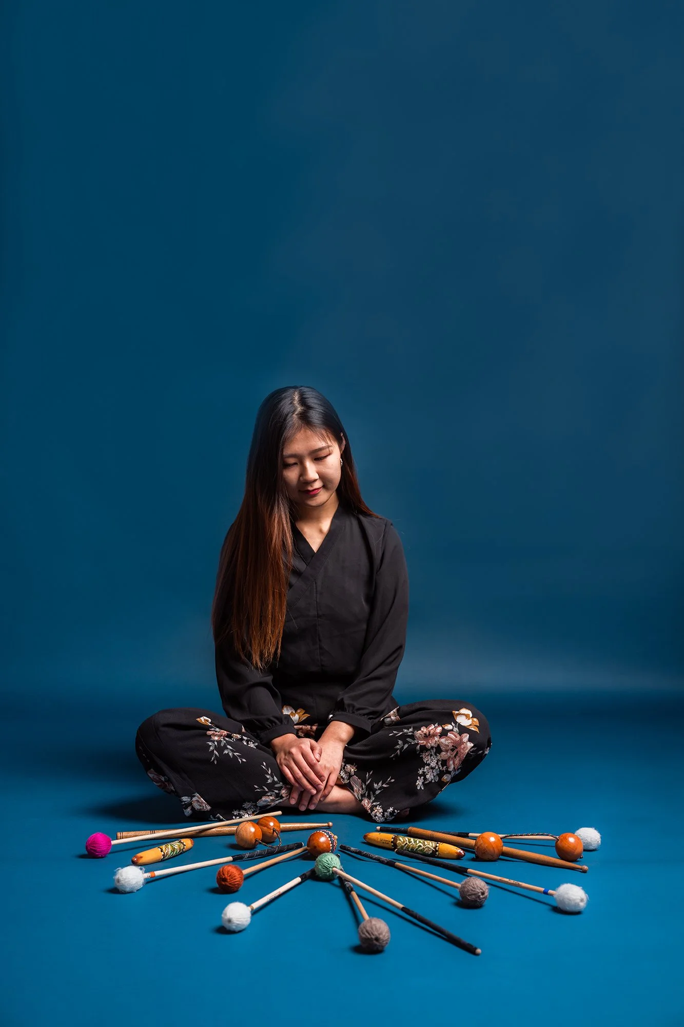 A young woman sitting cross-legged on the floor with her eyes closed, wearing a black kimono and floral pants, surrounded by colorful mallets and balls used for playing a traditional Japanese percussion instrument.