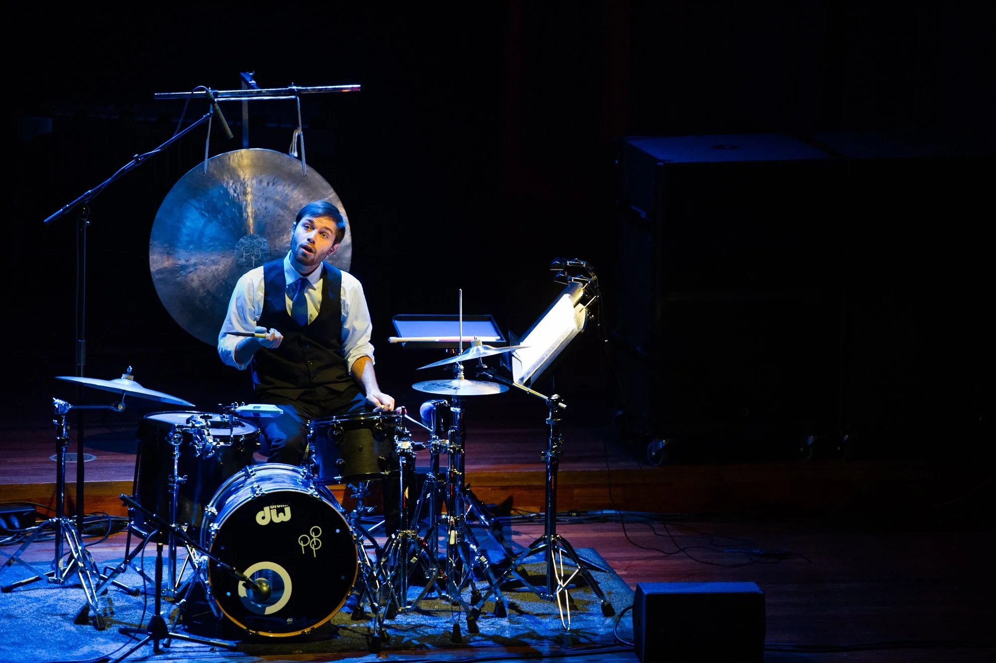 Dan DeSimone playing a setup piece during the final of the Tromp Percussion Eindhoven 2016 competition in formal attire sitting behind a drum set on a stage, with a large gong mounted behind him, illuminated by stage lights.