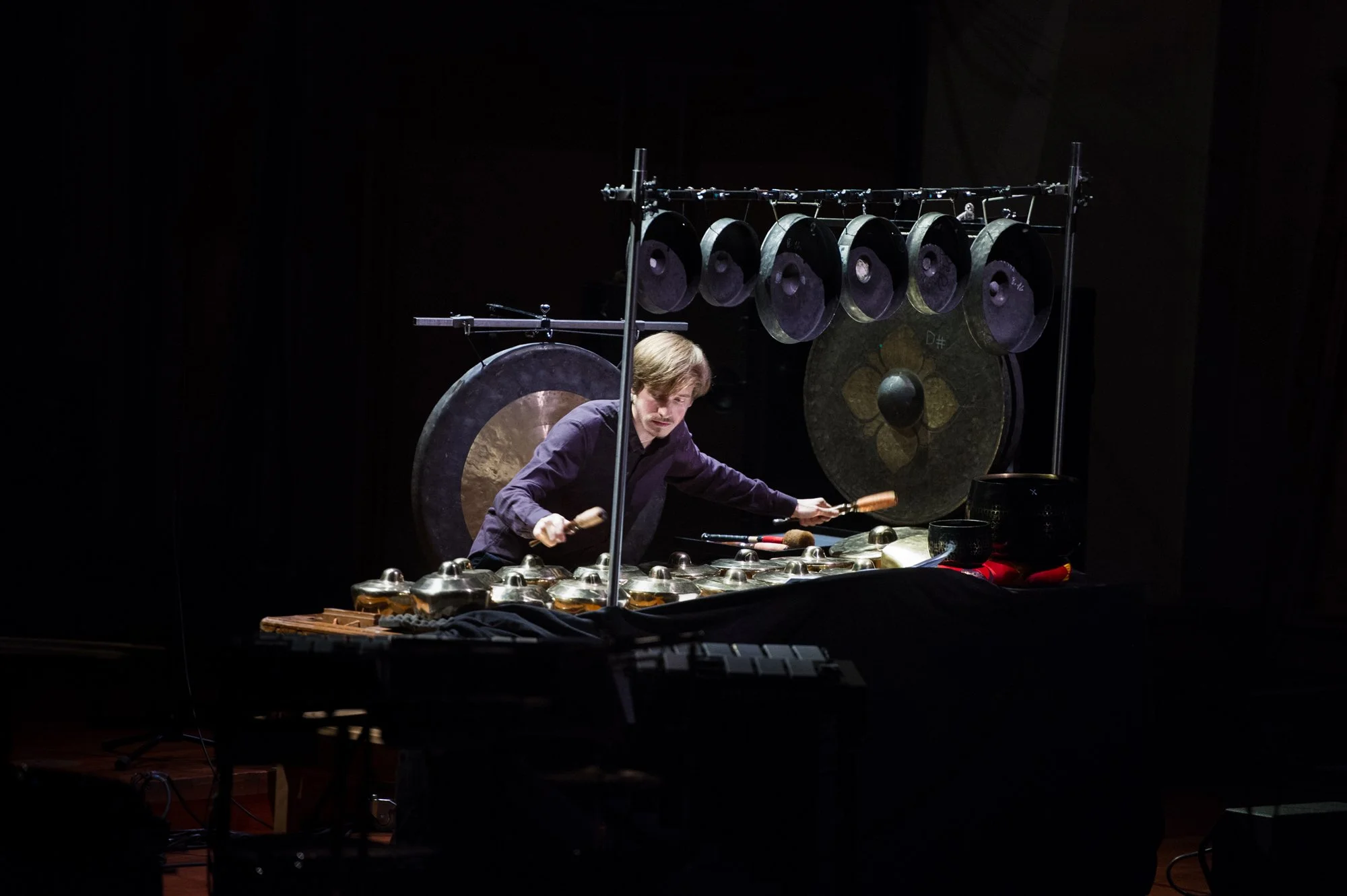 Sven Hoscheit playing a setup piece during the final of the Tromp Percussion Eindhoven 2016 competition with percussion instruments including gongs and bells on a dark stage.
