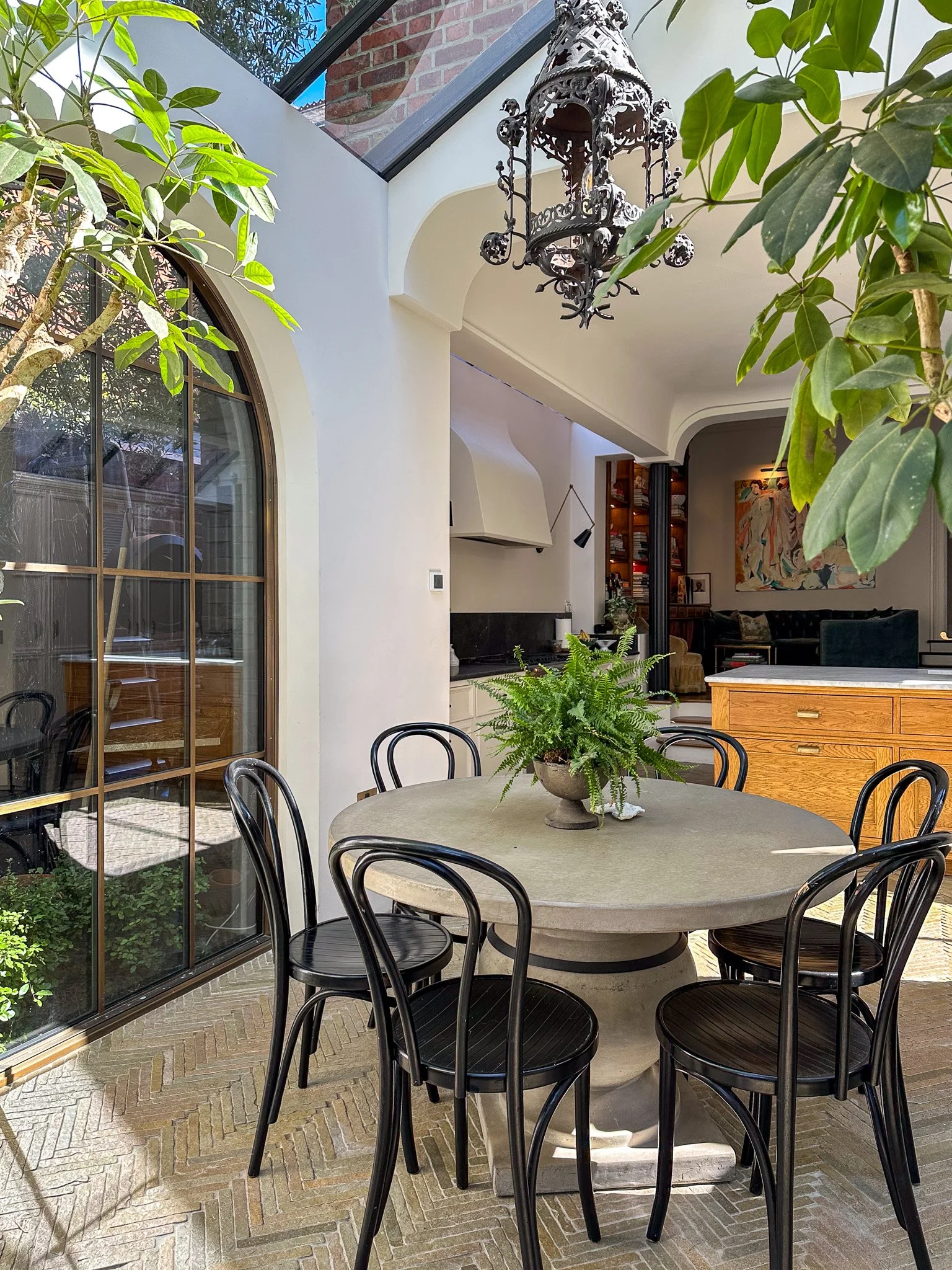 Indoor patio with a round table, black chairs, potted fern, large arched window, hanging lantern, interior bookshelves, and abstract painting in the background.