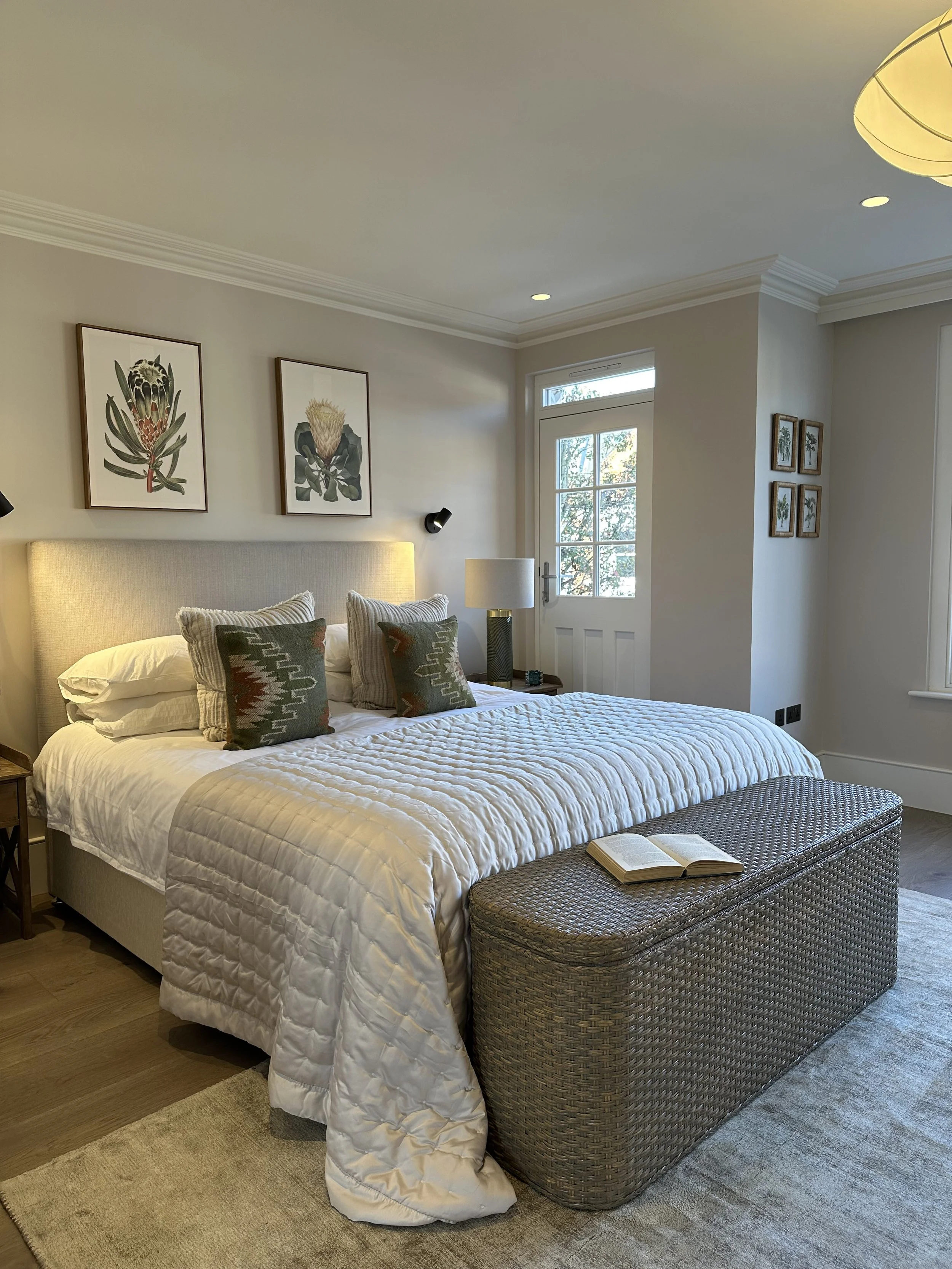 A cozy bedroom with a bed, decorative pillows, a woven bench with an open book, framed botanical art above, and a door leading outside, lit by natural light from a window.