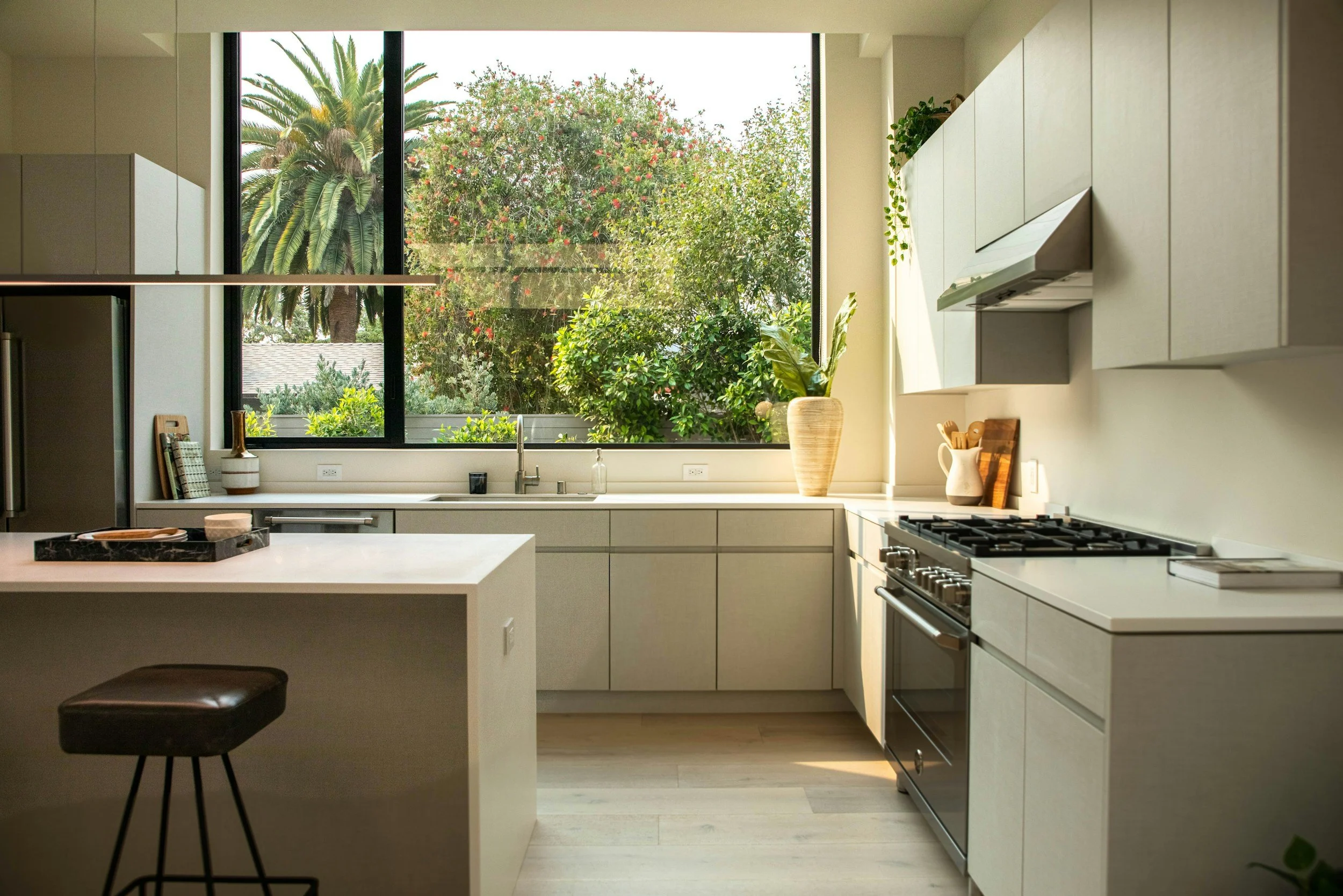 Modern kitchen with white cabinets, a large window showing trees and palm trees outside, a countertop with a potted plant, and a stove.