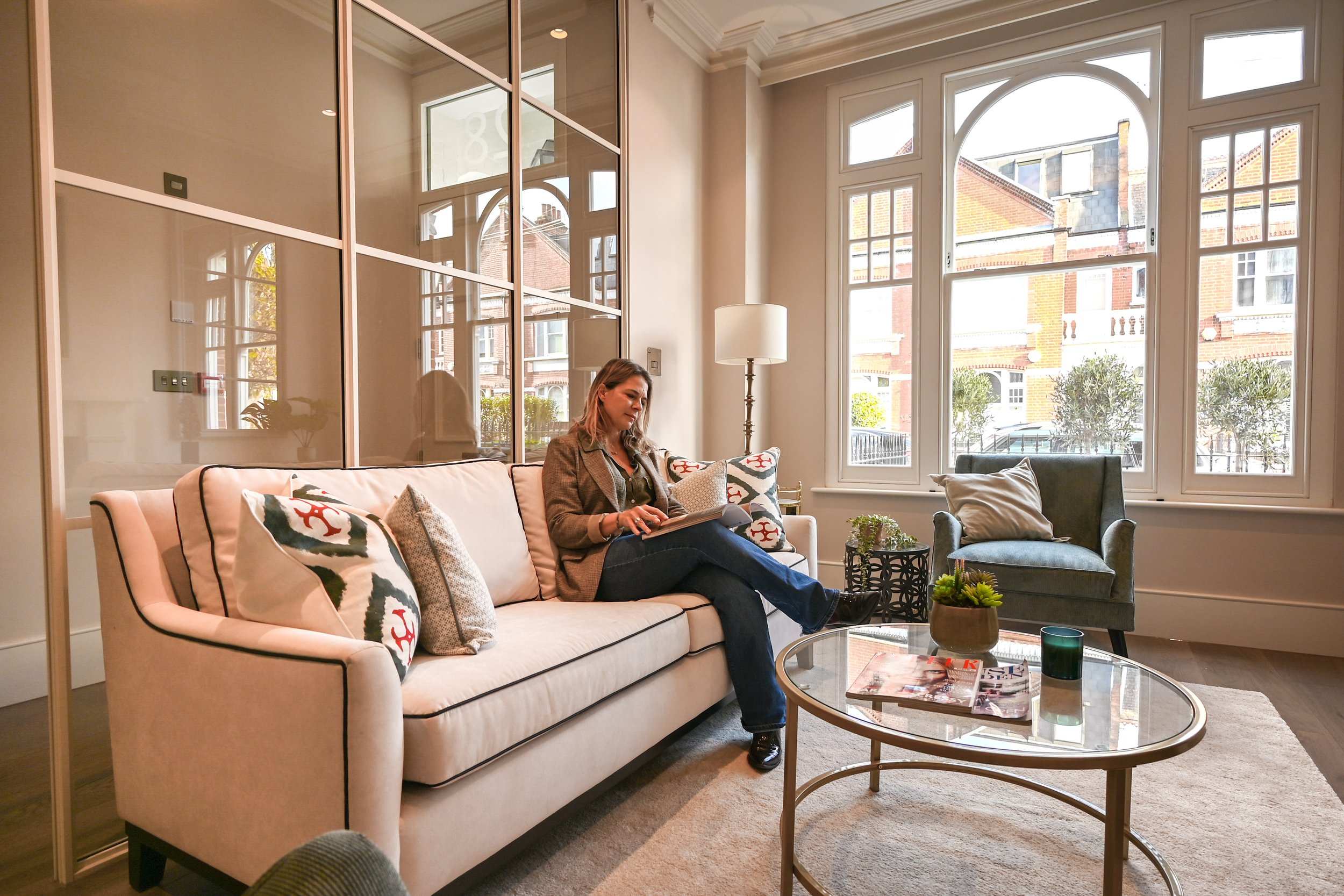 A woman sitting on a beige sofa reading a magazine in a well-lit living room with large windows, decorative pillows, a glass coffee table, and a gray armchair.