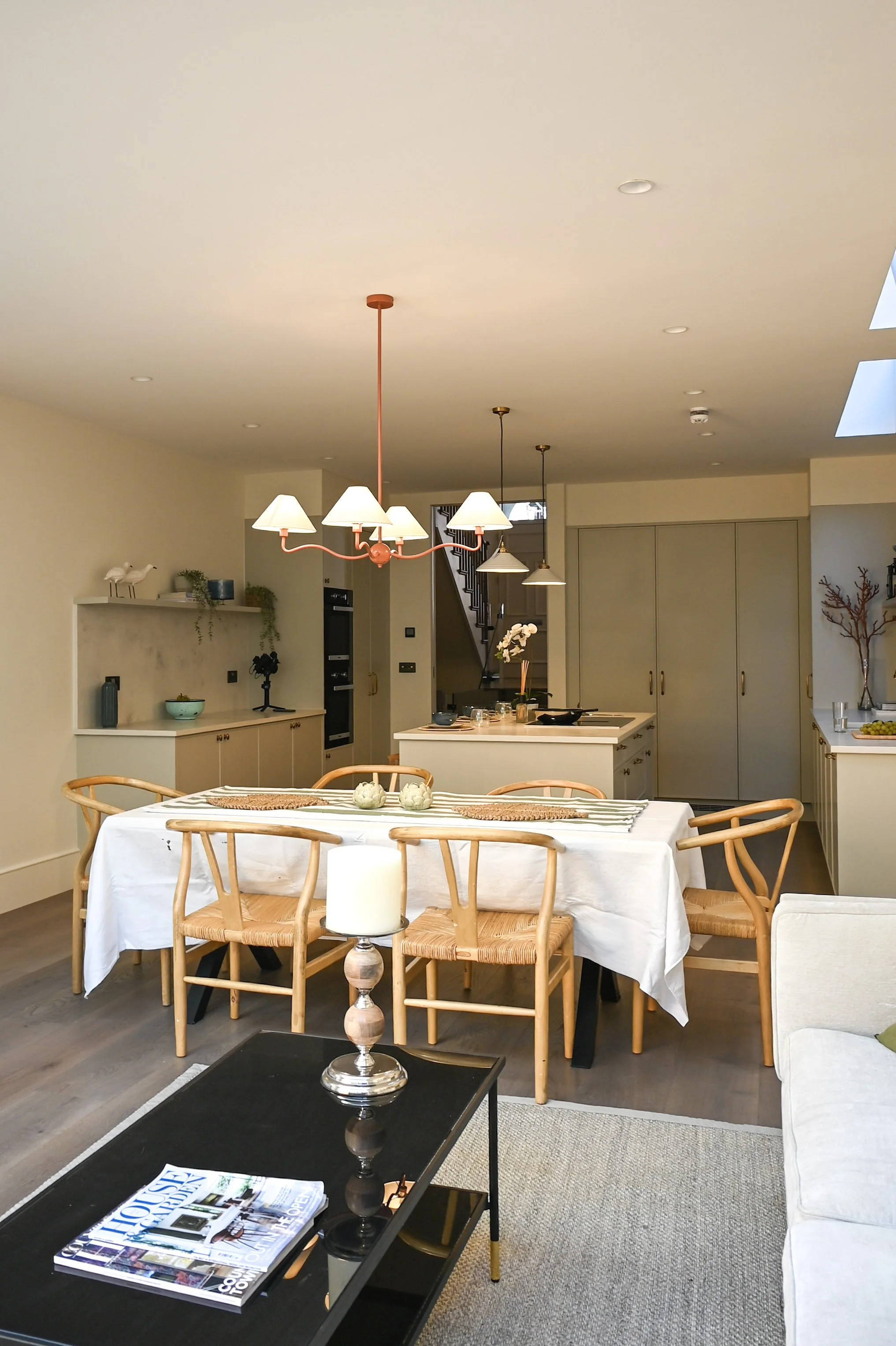 Open-concept dining and kitchen area with a white table, tan wooden chairs, and a black coffee table near a beige couch. The kitchen features a white island with a orchid plant, pendant lights, and neutral cabinets. Decor includes a lamp, dishes, and wall art.