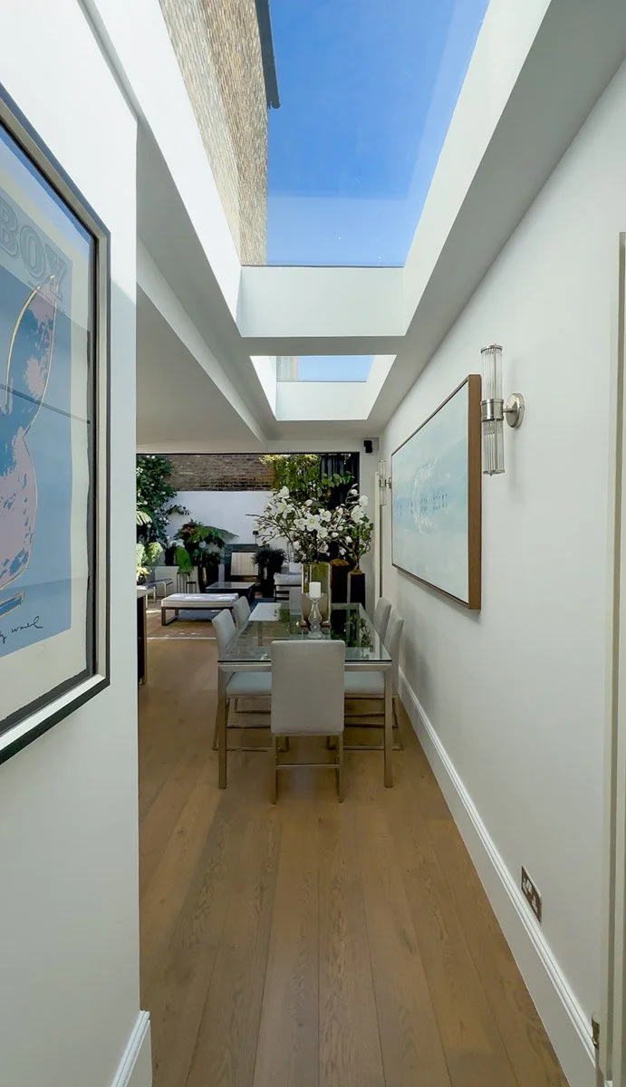 Modern dining room with skylights, wooden flooring, a glass table, white chairs, floral decor, and an outdoor patio area visible through the skylights.