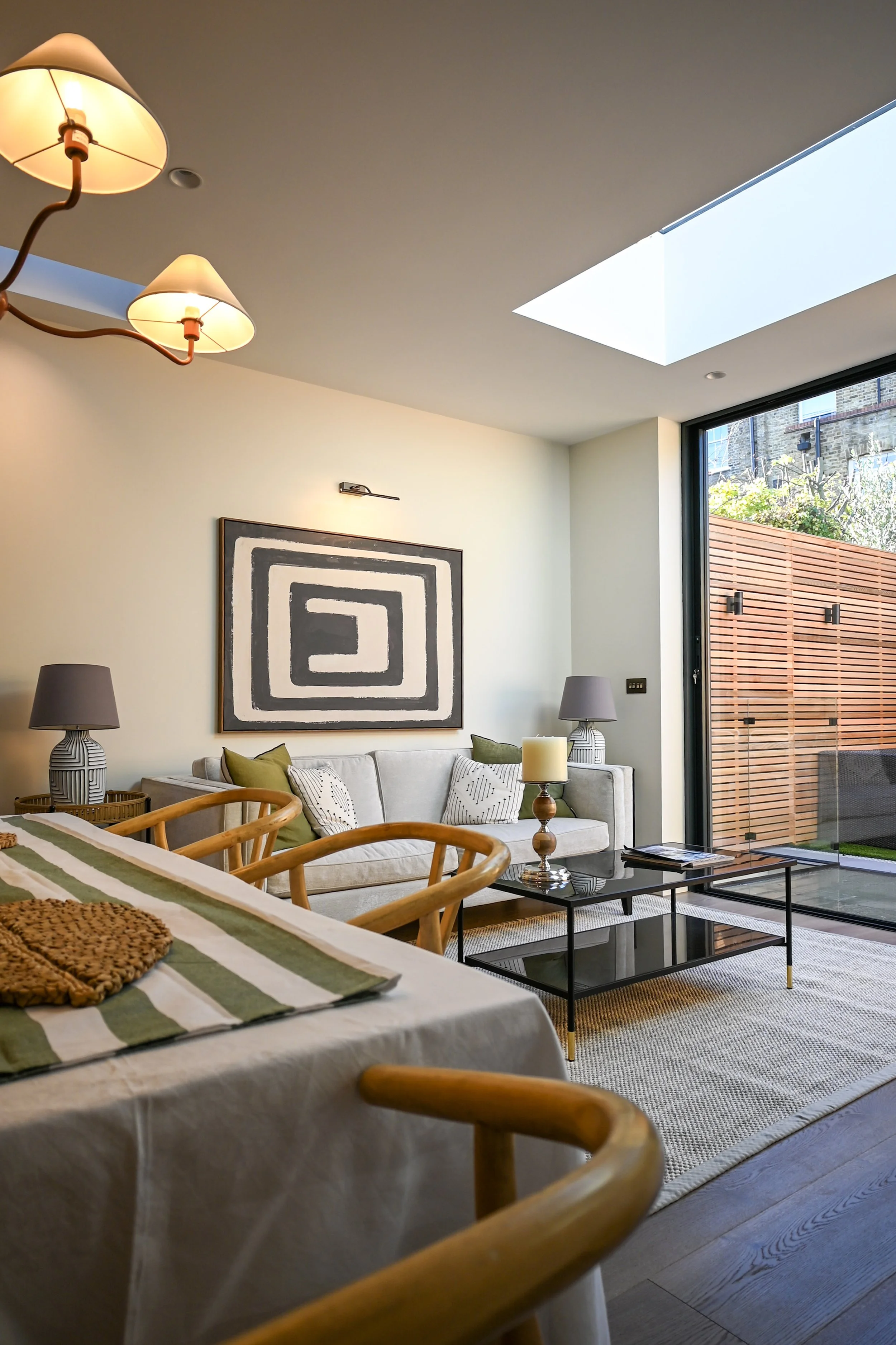 Modern living room with a coffee table, white sofa with pillows, large glass sliding door with wooden fence outside, abstract wall art, and skylight.