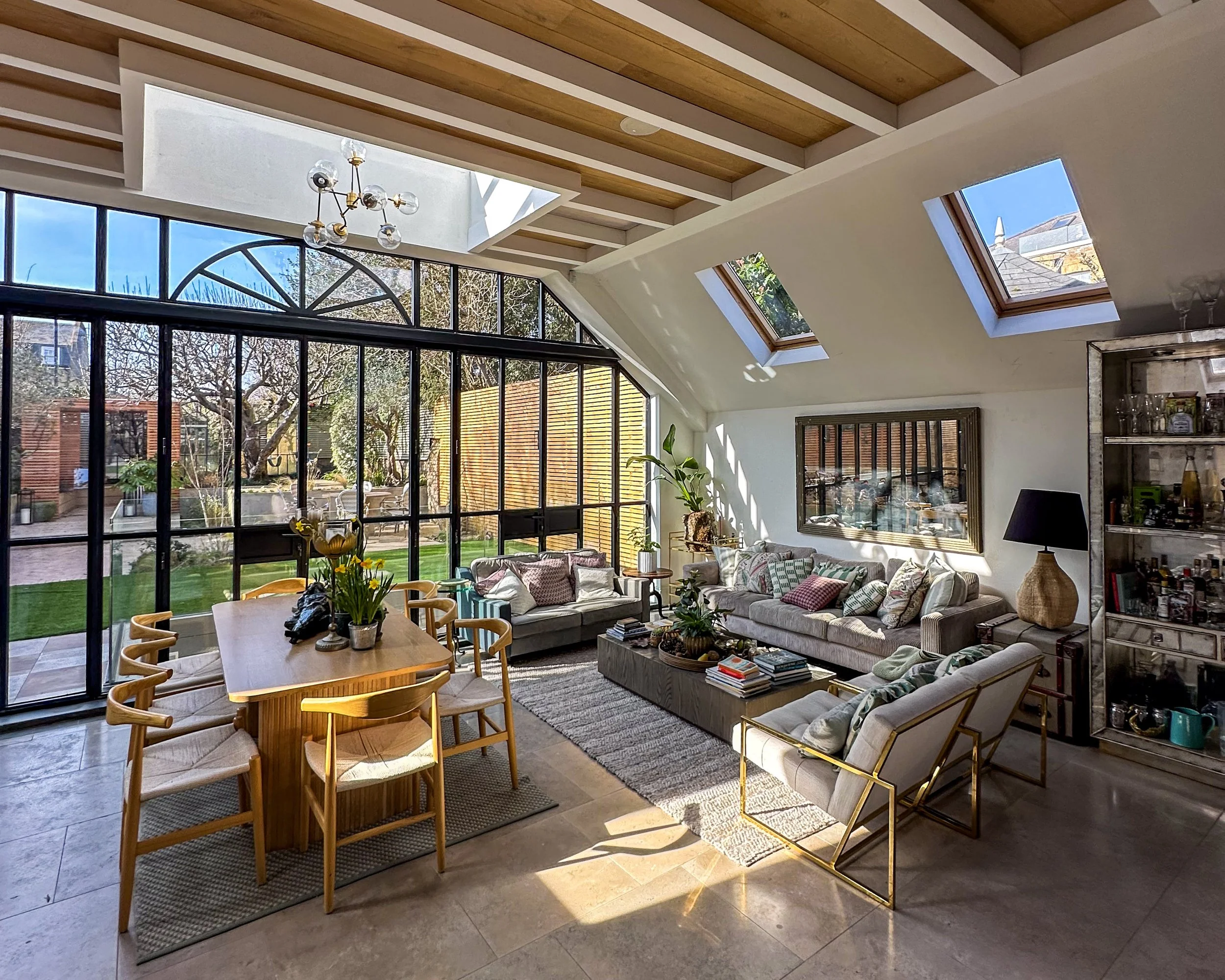 Bright living room with large glass windows and skylights, featuring a wooden dining table with six chairs, a gray sofa, armchairs, a coffee table with books, a side table, a tall plant, and a bookshelf against the wall. Outdoor garden visible through windows.