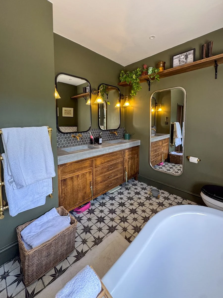 Bathroom with green walls, a white bathtub, wooden vanity with two sinks, black hexagon tile backsplash, and gold fixtures. Towel rack with white towels, a mirror, a large wall mirror, and a wooden shelf with decorative items and a plant.