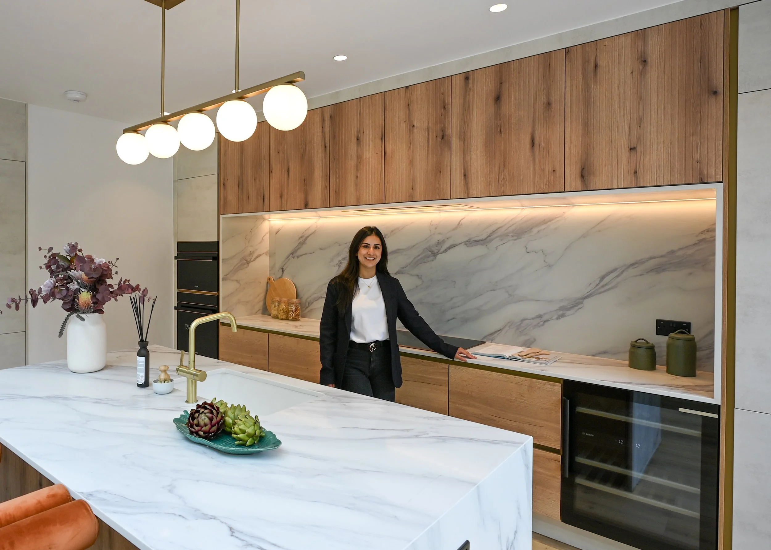 A woman standing in a modern kitchen with wooden cabinets, a marble backsplash, and a marble kitchen island with a gold faucet. The kitchen features decorative items and lighting, and the woman is smiling at the camera.